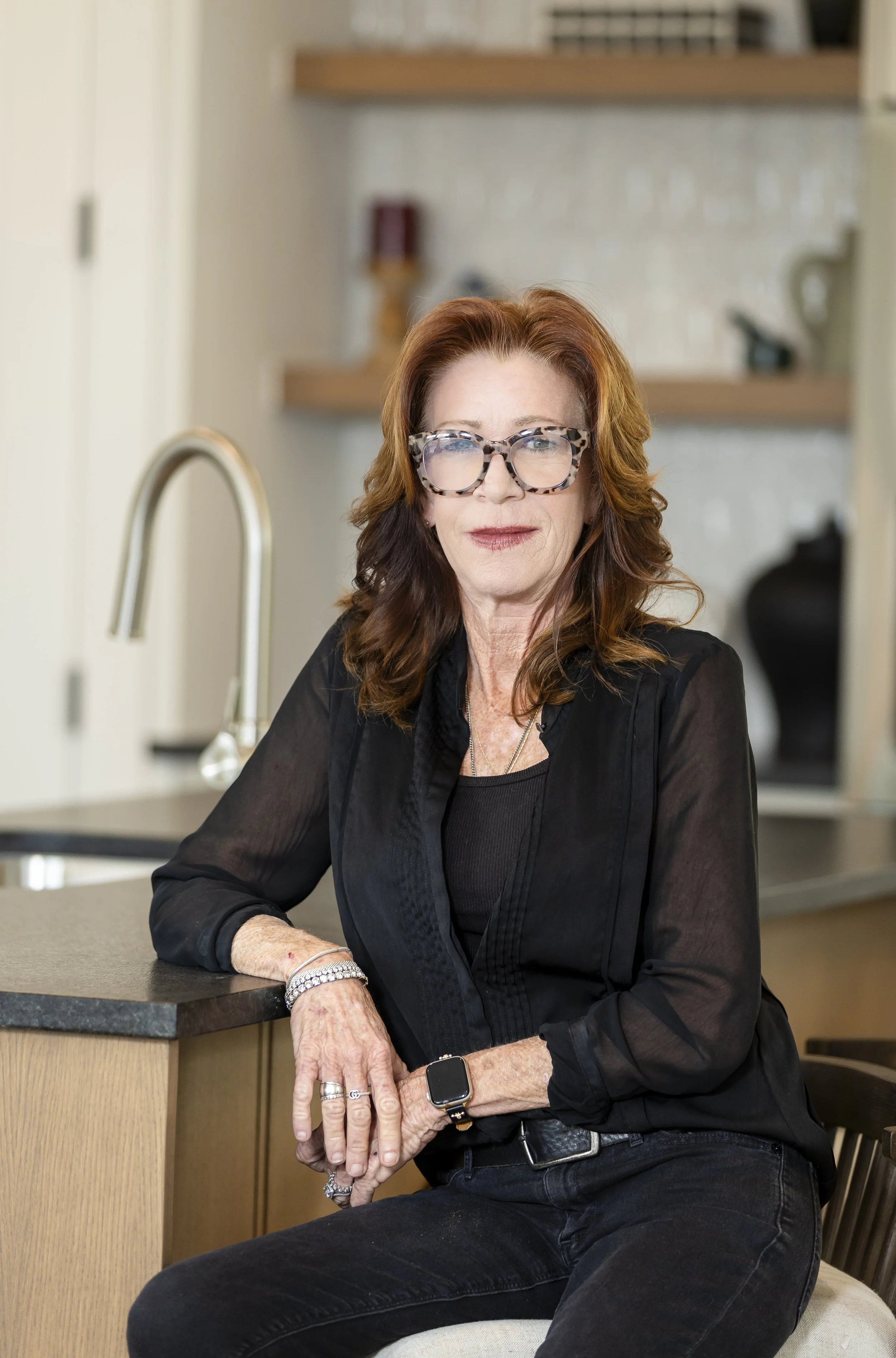 A woman with red hair wearing glasses and a black top, sitting at a kitchen counter with a faucet, in a modern kitchen.