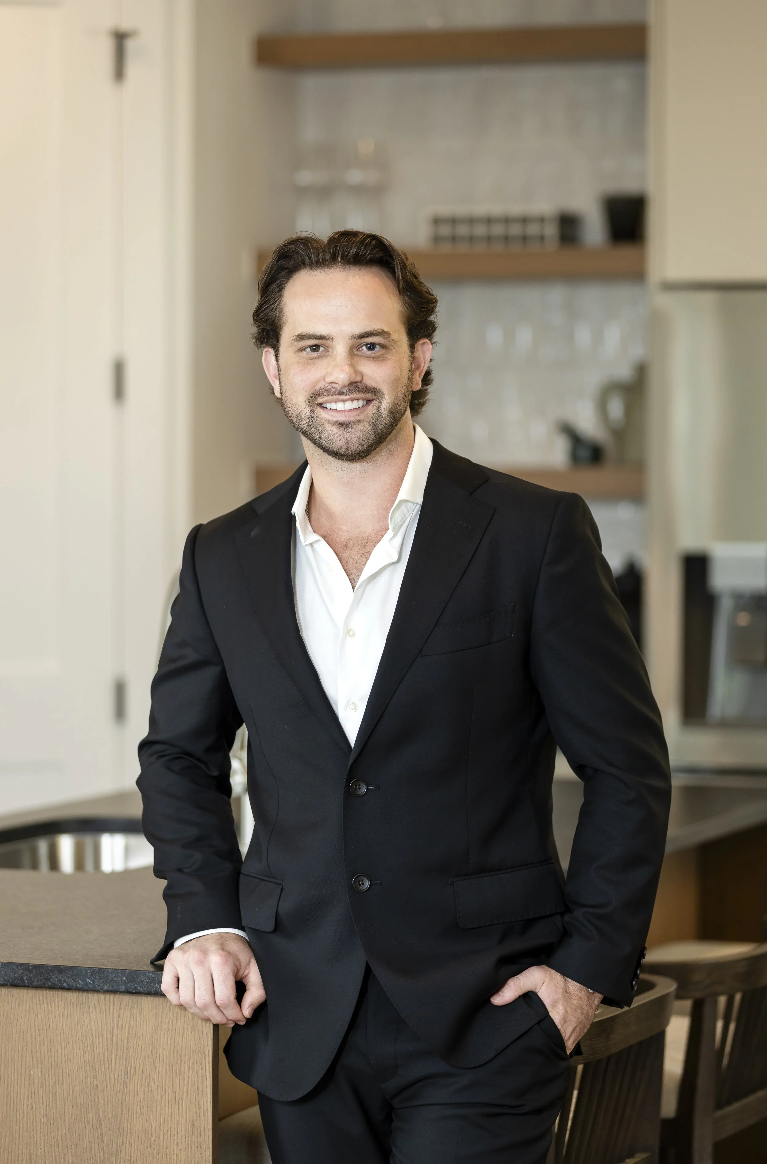 Man in black suit with white shirt smiling and leaning against a kitchen counter.