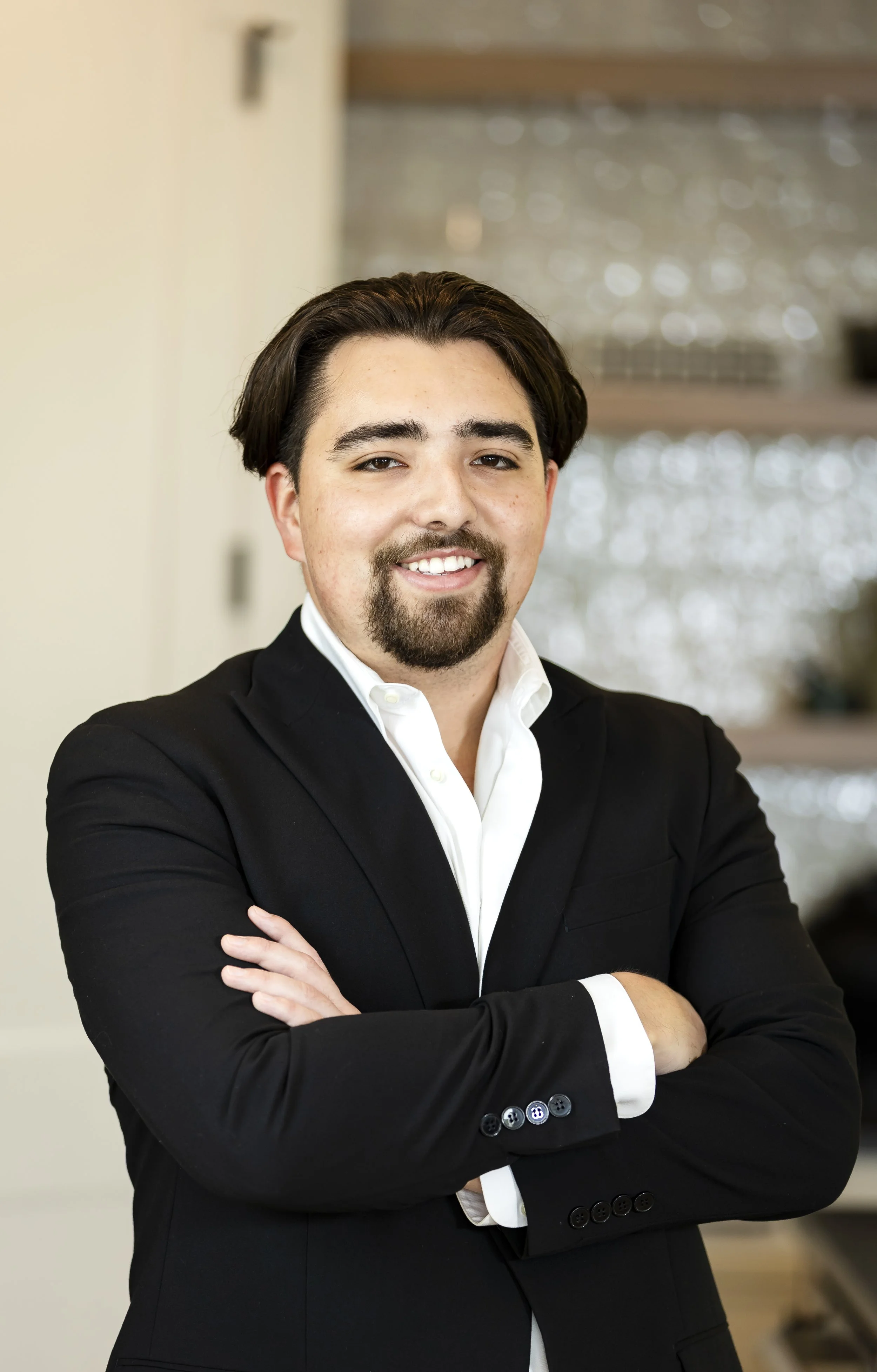A young man in a black suit and white shirt standing with arms crossed and smiling, in an indoor setting with a blurred background.
