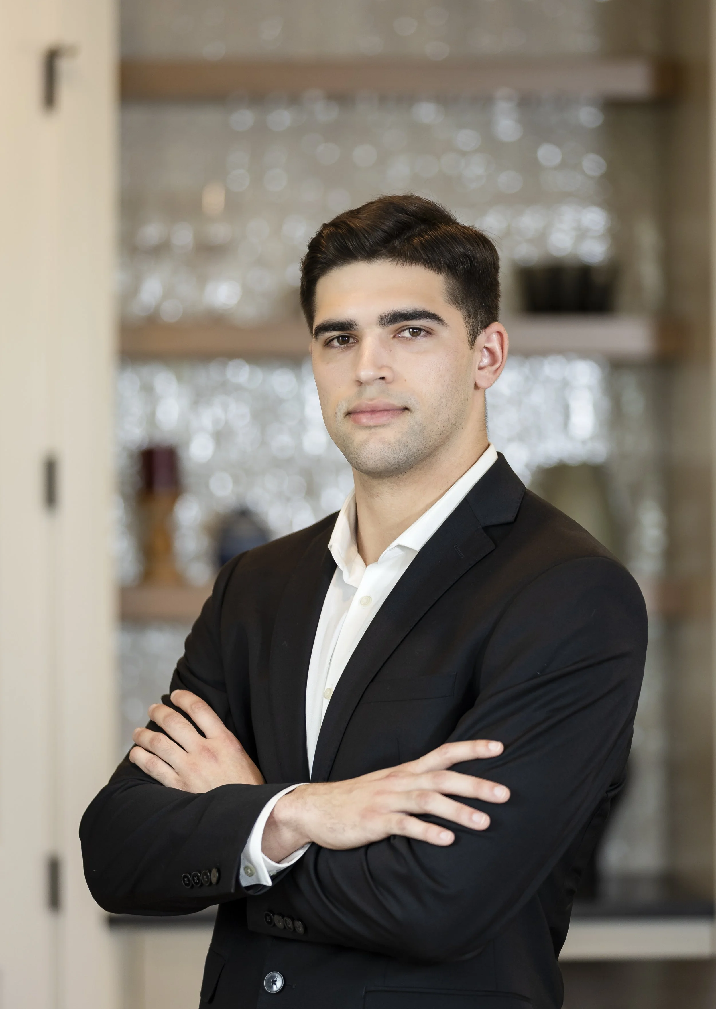 A young man wearing a black suit with a white shirt, standing with crossed arms in front of a blurred background.