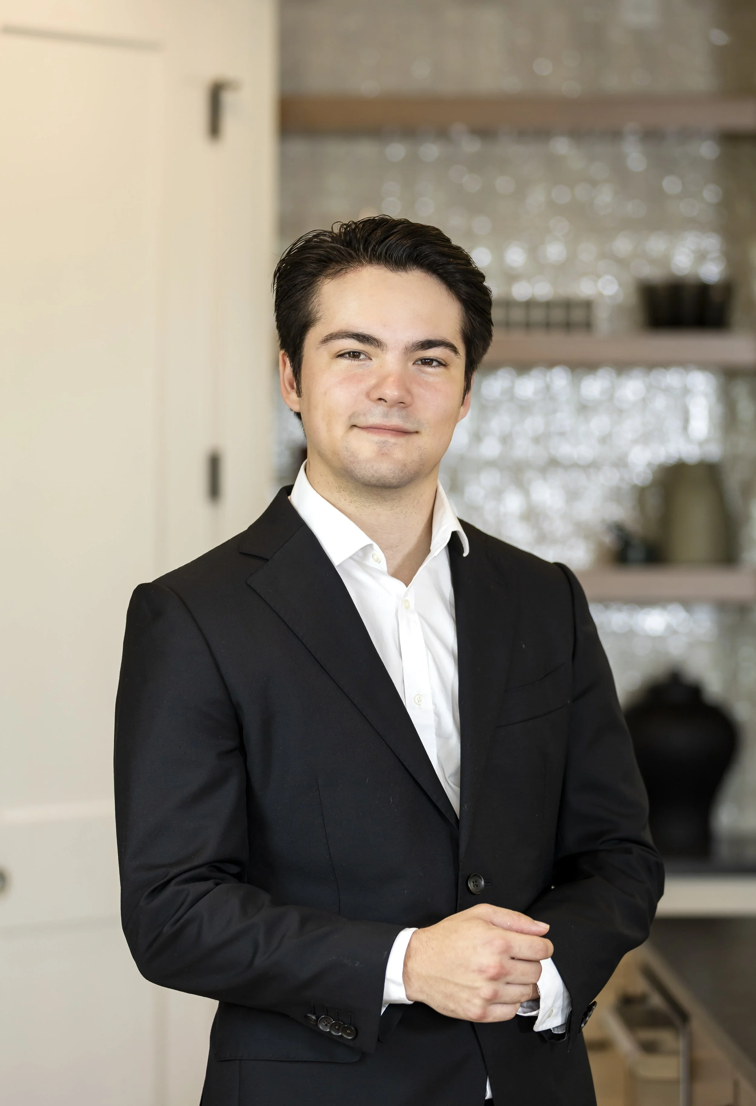 A young man dressed in formal attire, wearing a black suit and white shirt, standing indoors with a kitchen in the background.