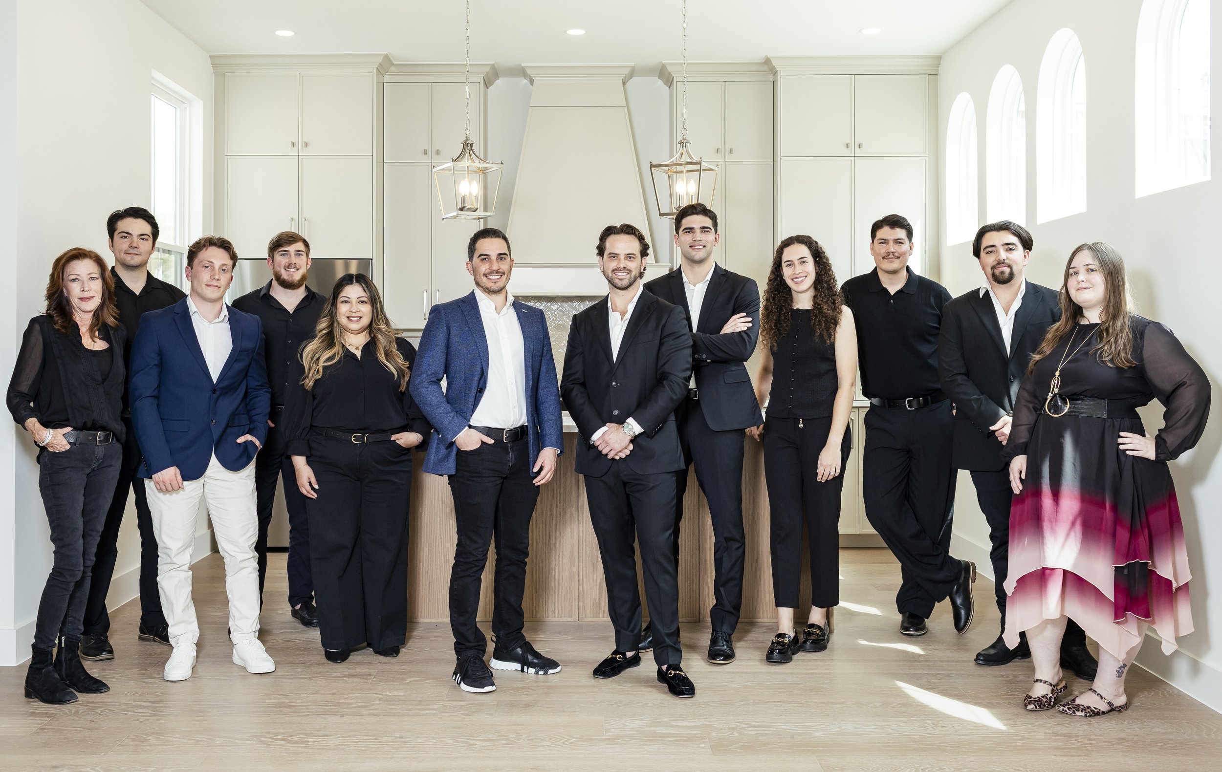 A group of 13 diverse professionals in business attire standing in a bright, modern kitchen with white cabinets and large windows.