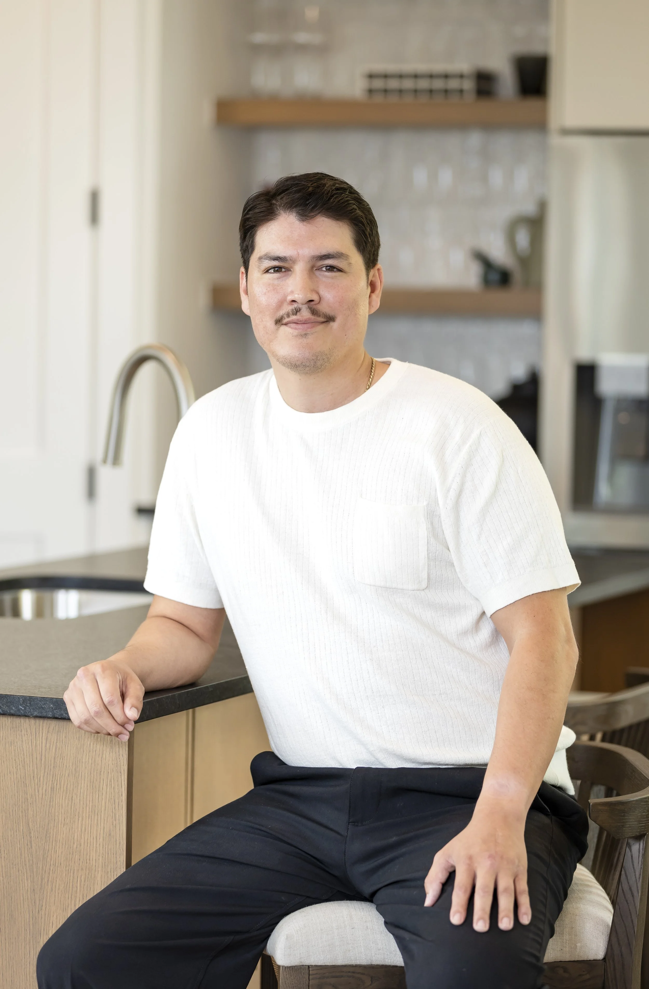 A man with dark hair and a mustache sitting on a chair in a modern kitchen, wearing a white short-sleeved shirt and black pants.