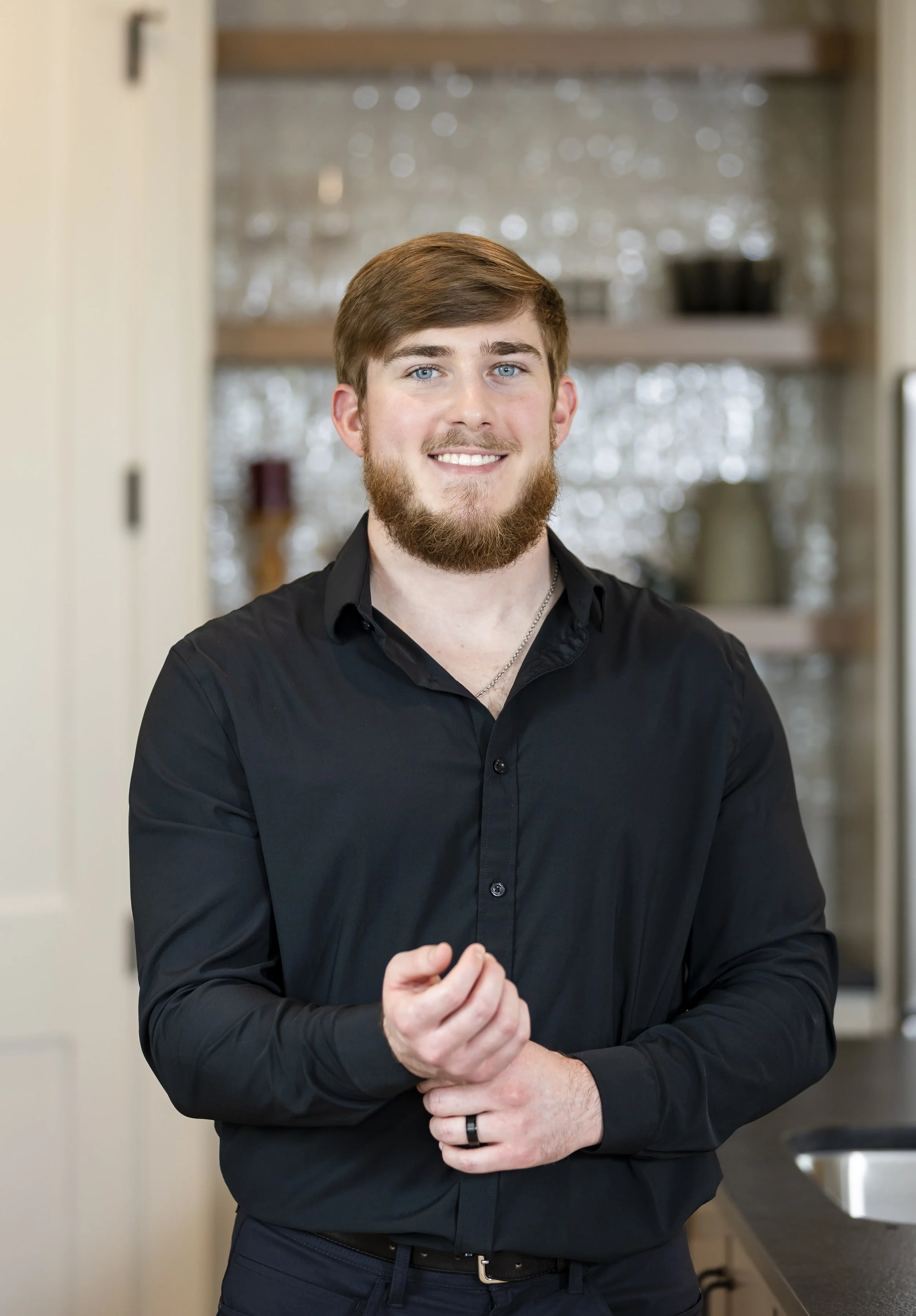 A smiling young man with a beard and brown hair, wearing a black dress shirt, standing in a kitchen with shelves and a backsplash visible in the background.