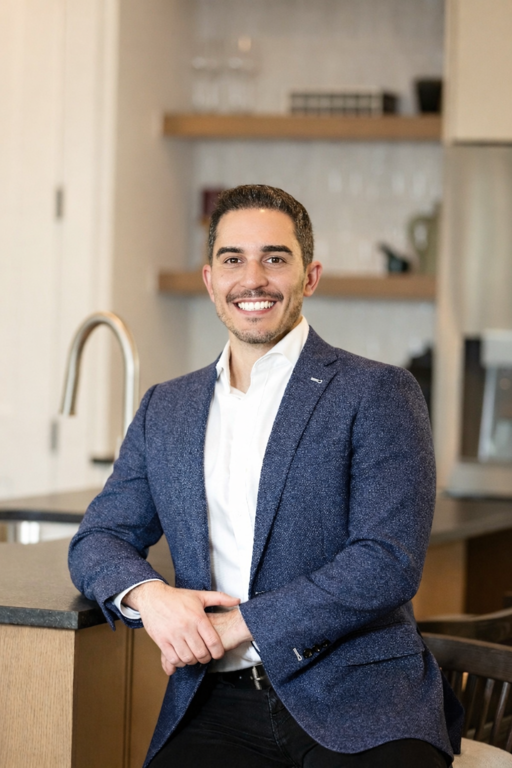 A man with dark hair, a beard, and light skin, smiling and wearing a white shirt and a dark blue blazer, standing in a modern kitchen with wooden shelves and a gray background.