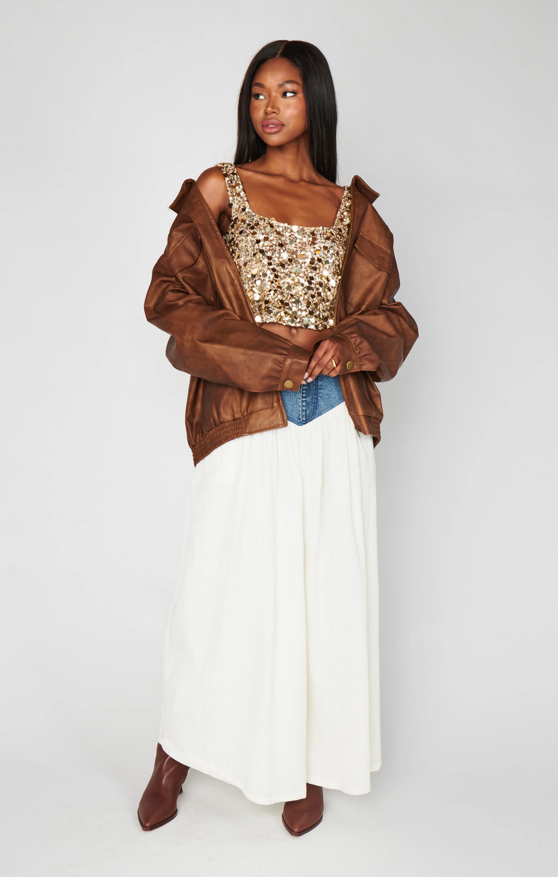 A woman dressed in a sparkly gold crop top, a brown leather jacket draped over her shoulders, a long white skirt, and brown boots, standing against a plain light gray background.