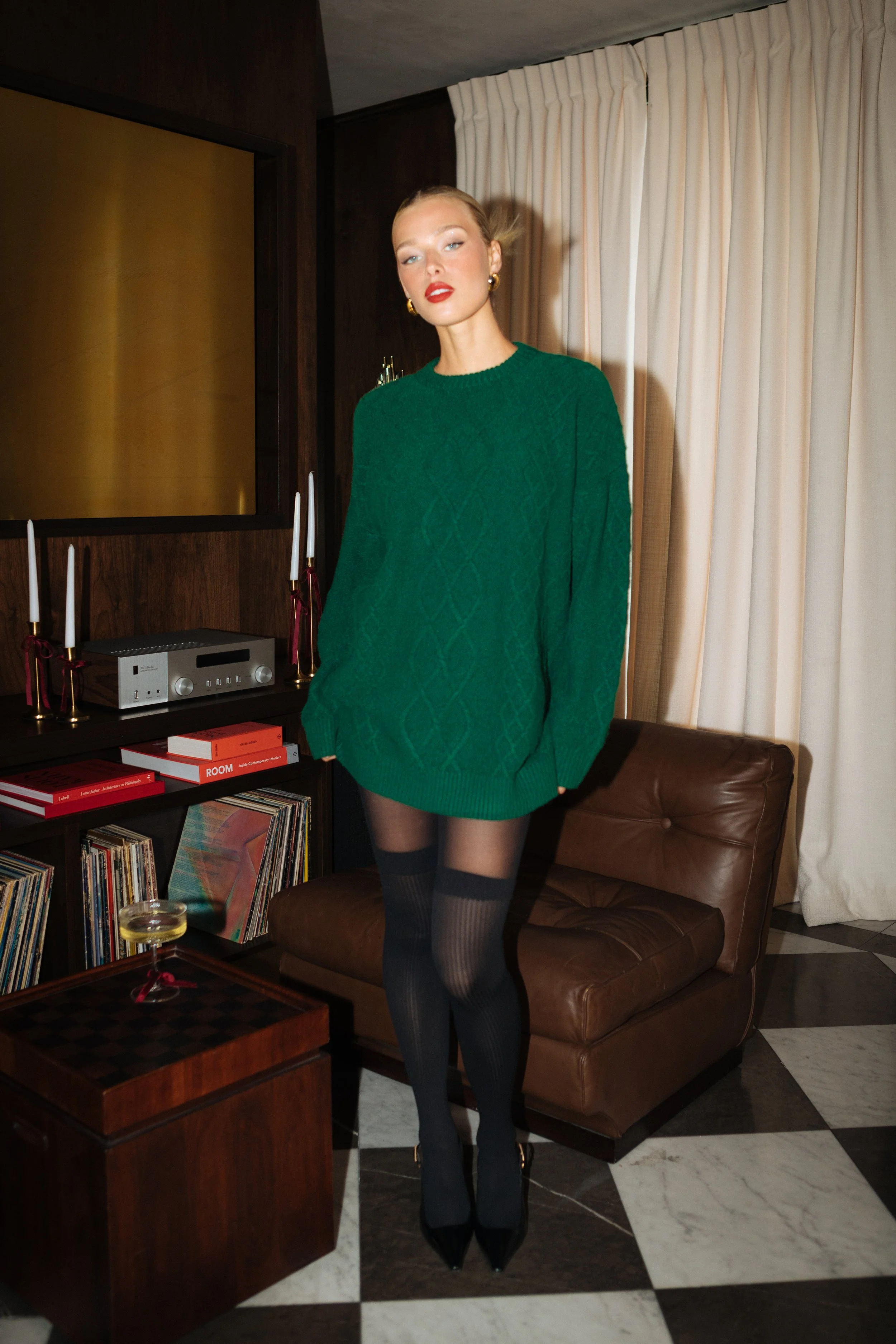 A woman standing in a living room with dark wood paneling, white curtains, a leather armchair, and a bookshelf with vinyl records. She is wearing a green oversized sweater, black sheer tights, and high heels.
