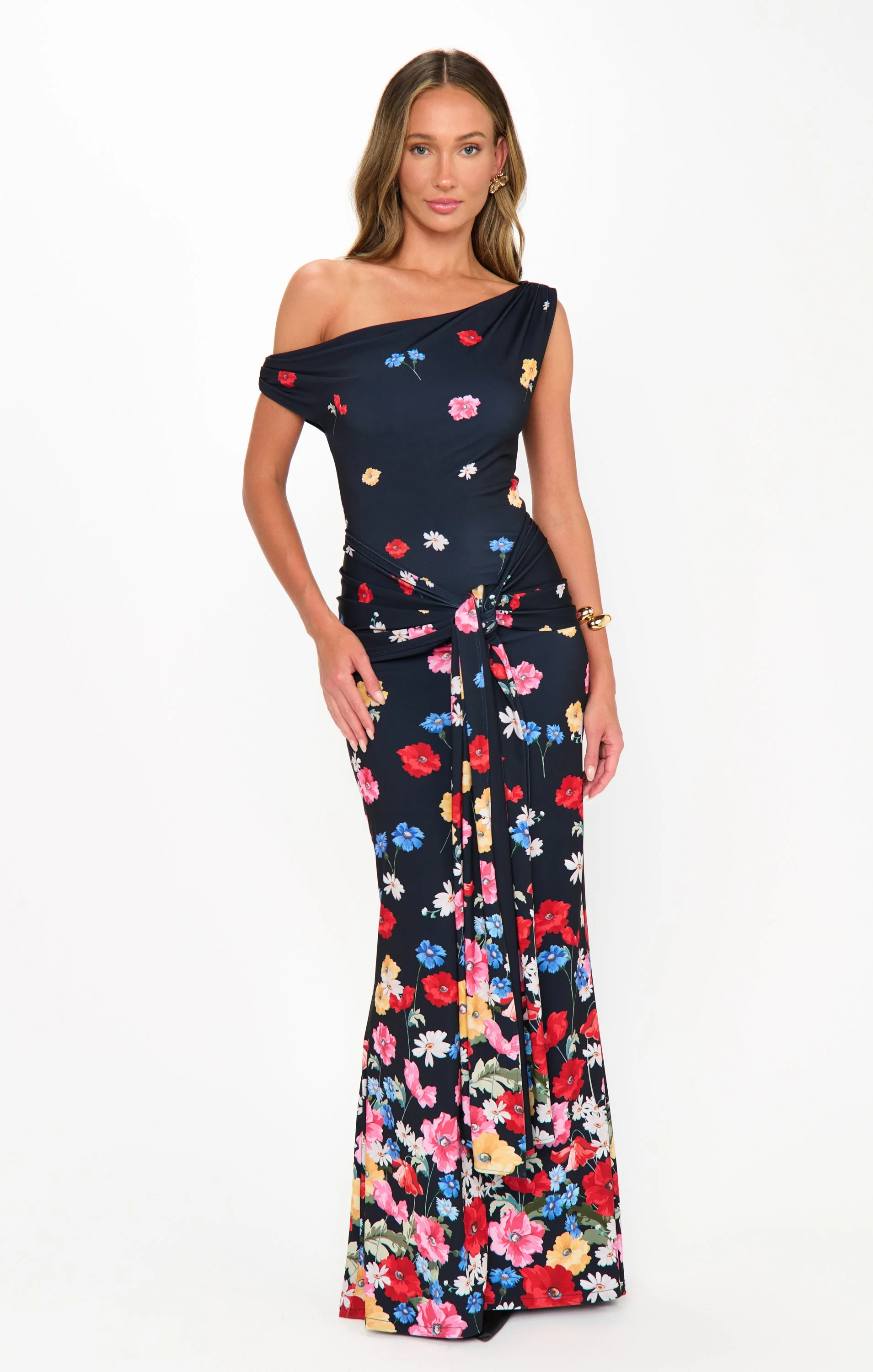 A woman wearing a navy off-the-shoulder jumpsuit with a colorful floral pattern, standing against a plain white background.