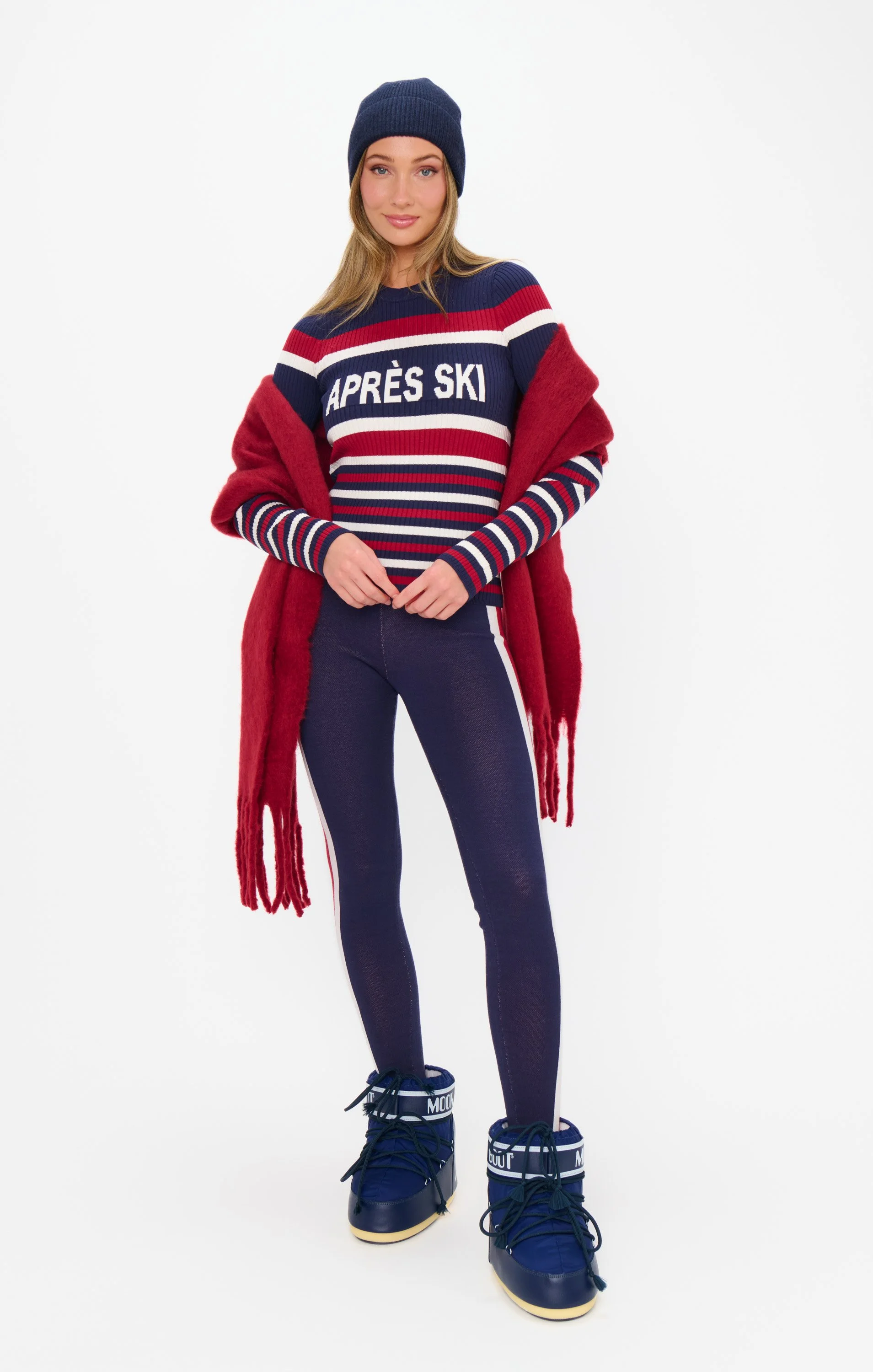 A young woman wearing a navy blue beanie, a red, white, and blue striped sweater with the words 'APRÈS SKI' printed on it, a red fuzzy cardigan, navy blue ski pants with white stripes, and blue snow boots with ankle straps.