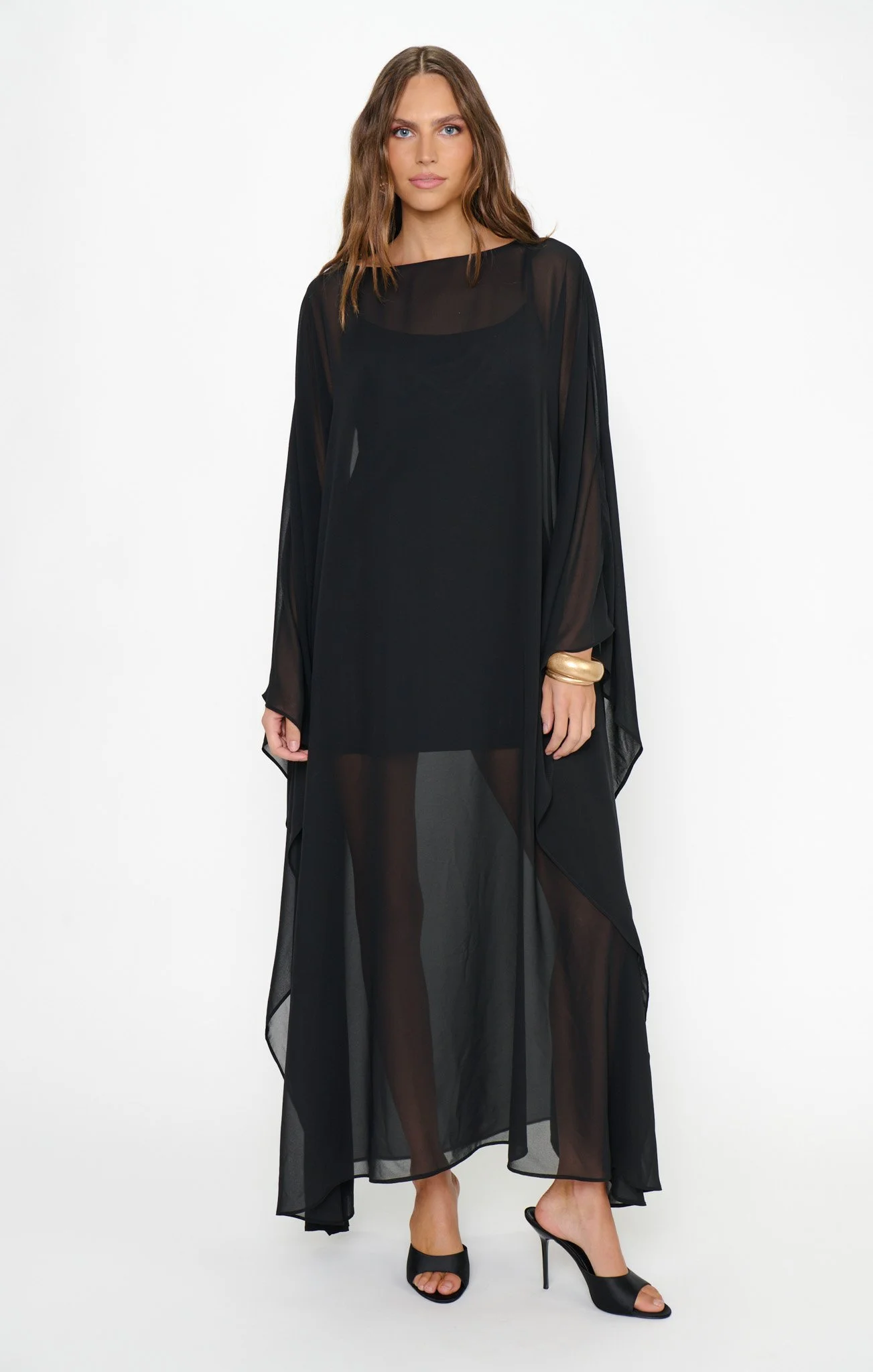 Woman wearing a black sheer dress with a slip underneath, black high-heeled sandals, and gold bangles on her wrist.