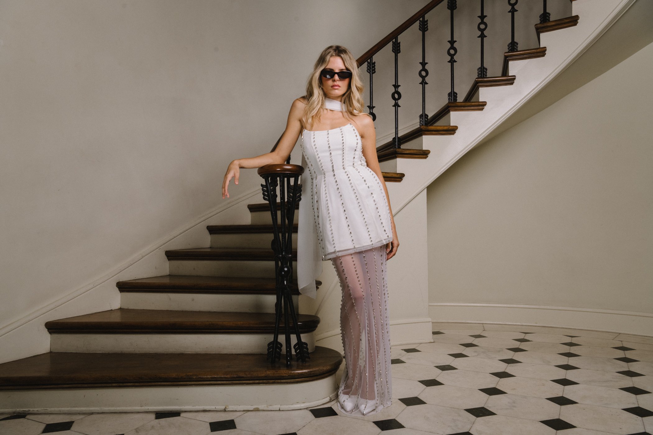 Fashion model wearing sunglasses, a white strapless dress with decorative eyelet embellishments, and sheer pants, leaning against a staircase railing in an elegant indoor setting.
