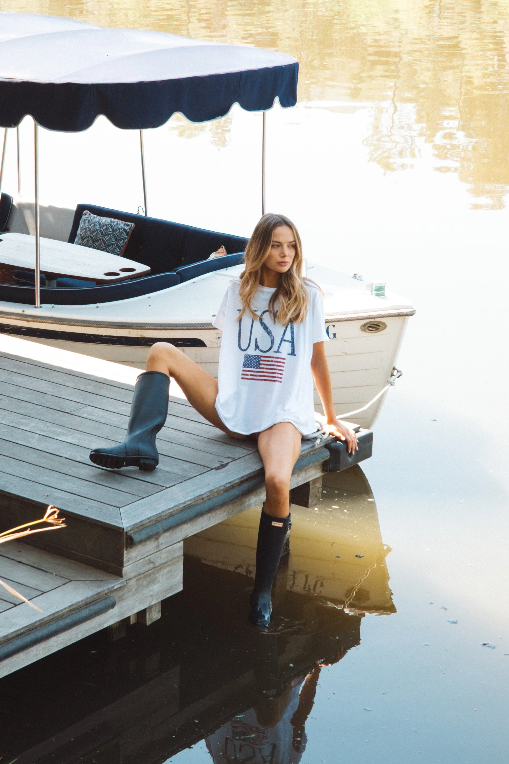 A woman sitting on a dock by a boat, wearing an American flag-themed t-shirt and black rain boots, with her feet in the water.