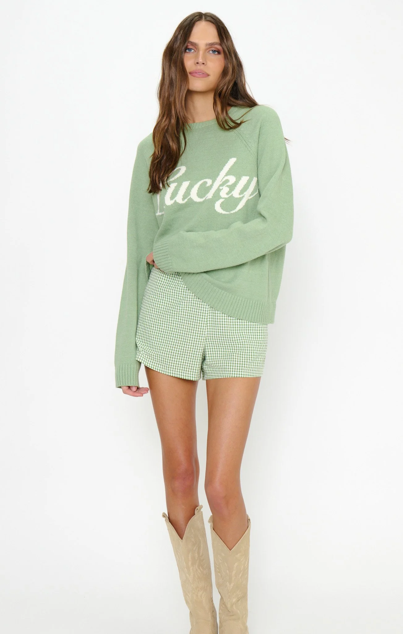A young woman with wavy brown hair wearing a light green sweater that says 'Lucky', matching green and white checked shorts, and beige cowboy boots.
