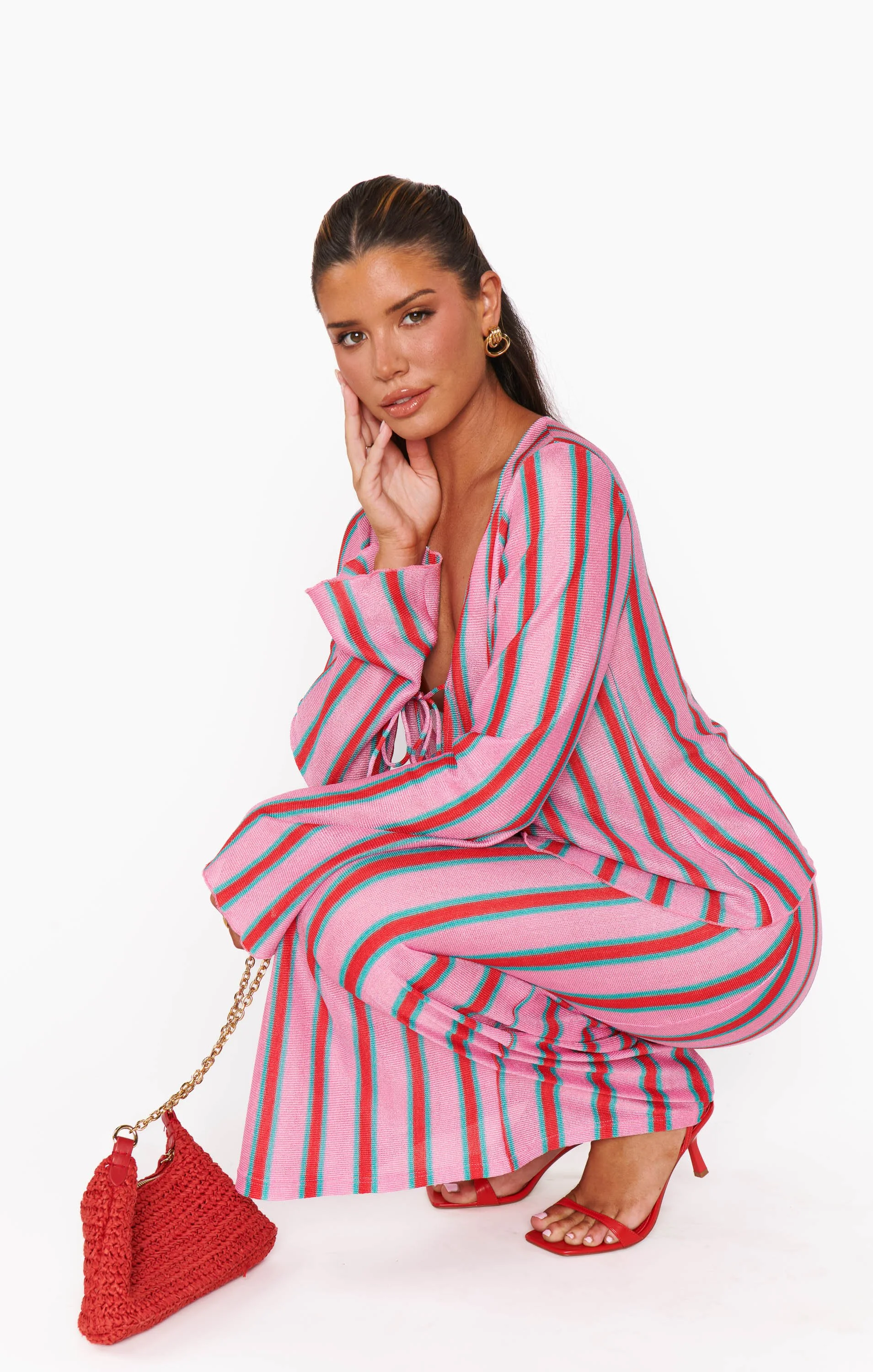 A woman in a pink and green striped matching dress and red heels squats against a plain white background with a red woven purse on a gold chain beside her.