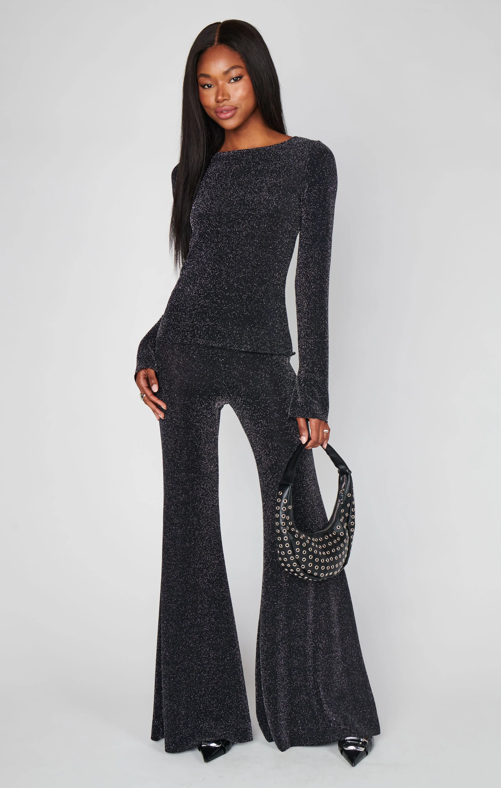 A woman with long black hair wearing a sparkly black long-sleeve top and matching wide-leg pants, holding a black handbag with silver eyelets, standing against a plain light gray background.