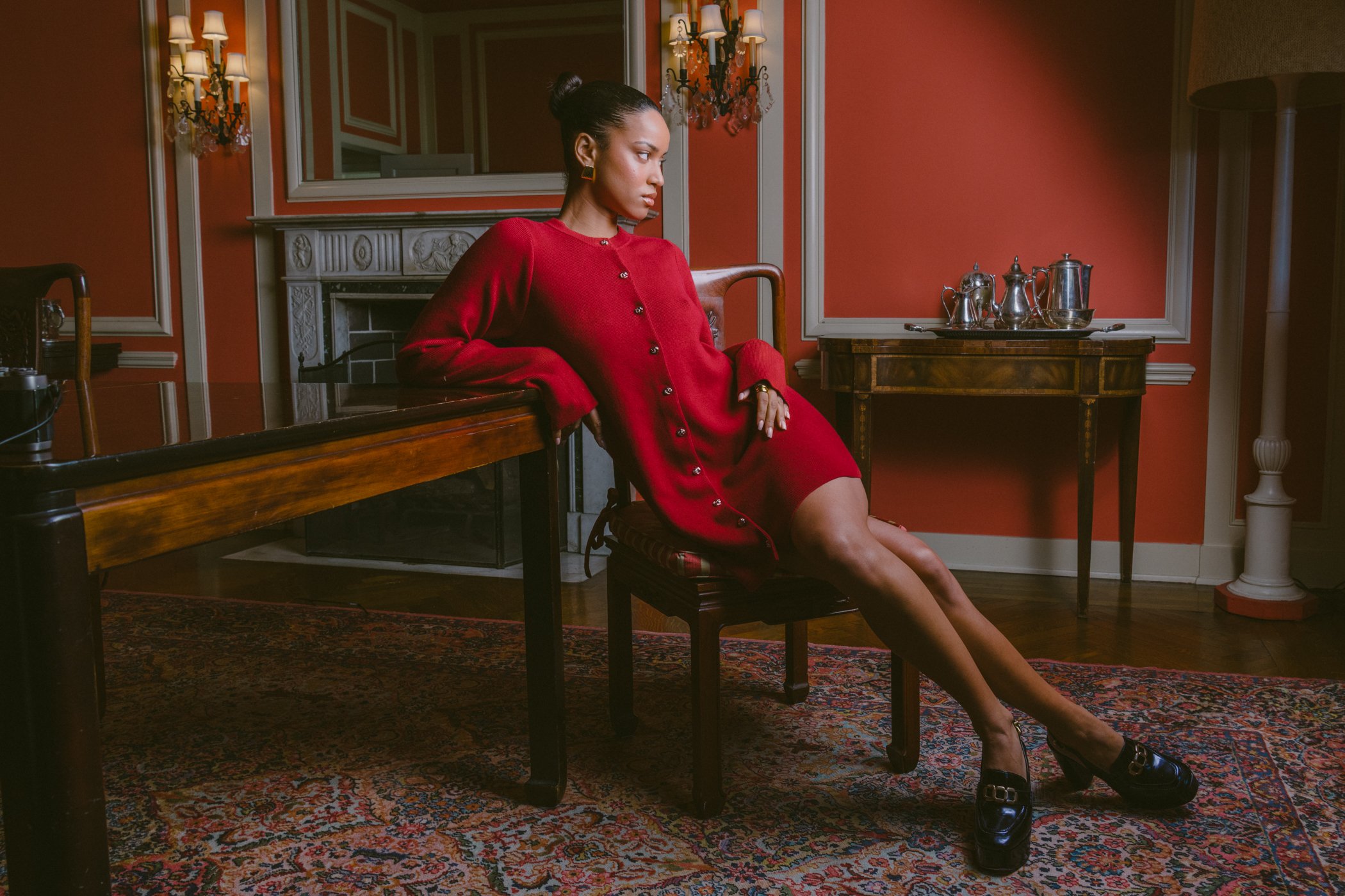 A woman in a red dress with gold buttons sitting on a chair in a room with orange walls, a fireplace, a mirror, a rug, and a table with silverware.