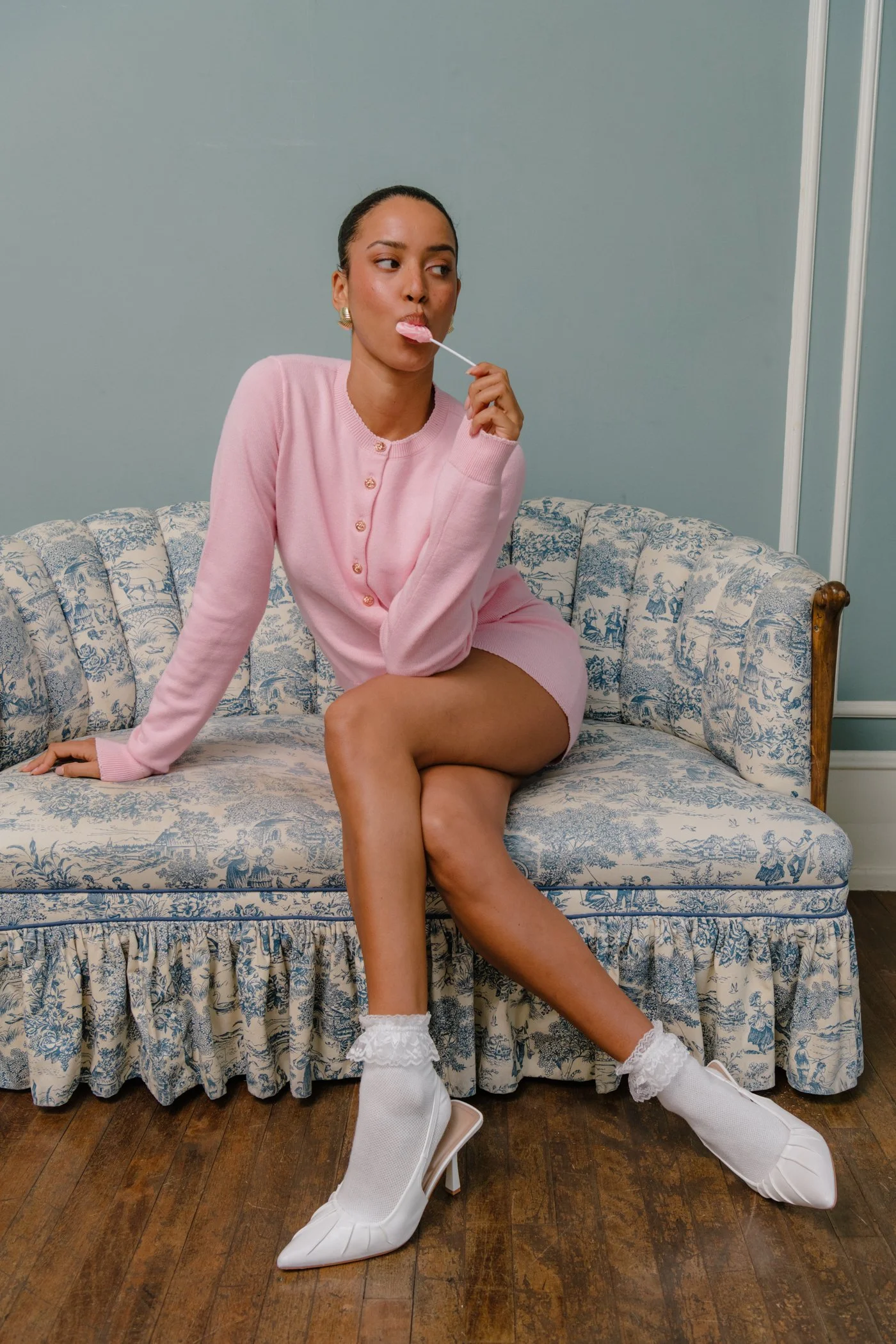A woman sitting on a vintage patterned sofa, wearing a pink cardigan and skirt, white lace socks, and white high-heeled shoes, licking a pink lollipop, with a neutral expression, against a plain blue wall.