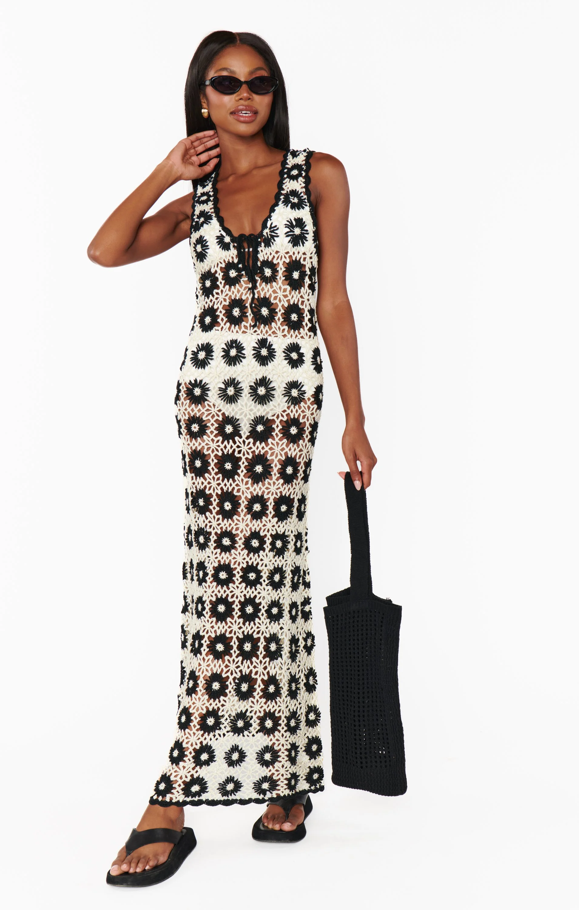 A woman in a long black and white floral crochet dress, black sunglasses, and black sandals holding a black woven bag, standing against a white background.