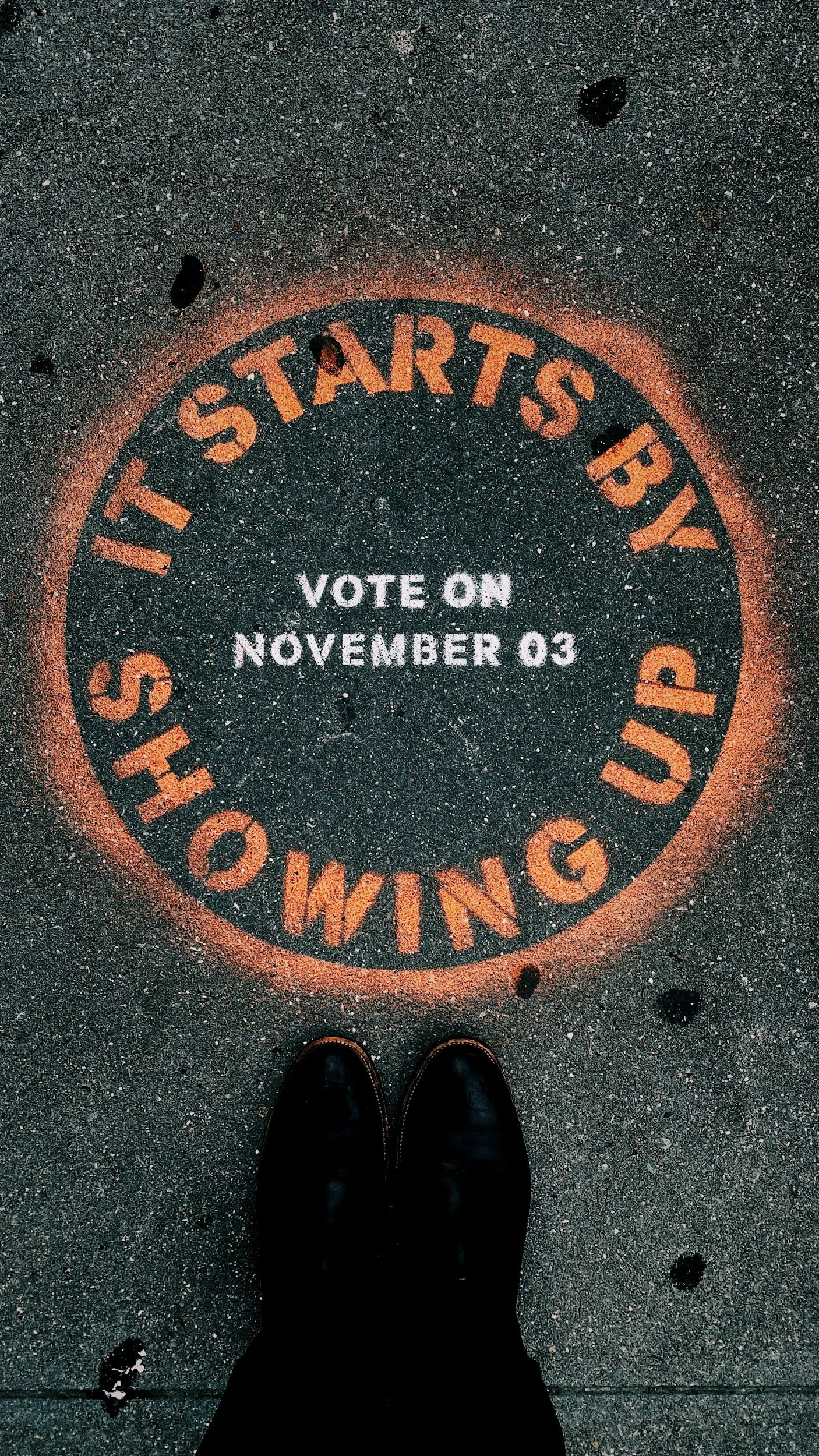 A circular sidewalk sign with the words 'STARTS BY SHOWING UP' in orange and white text, with steps to vote on November 3rd inside the circle. The photo is taken from a person's perspective, showing black shoes at the bottom.