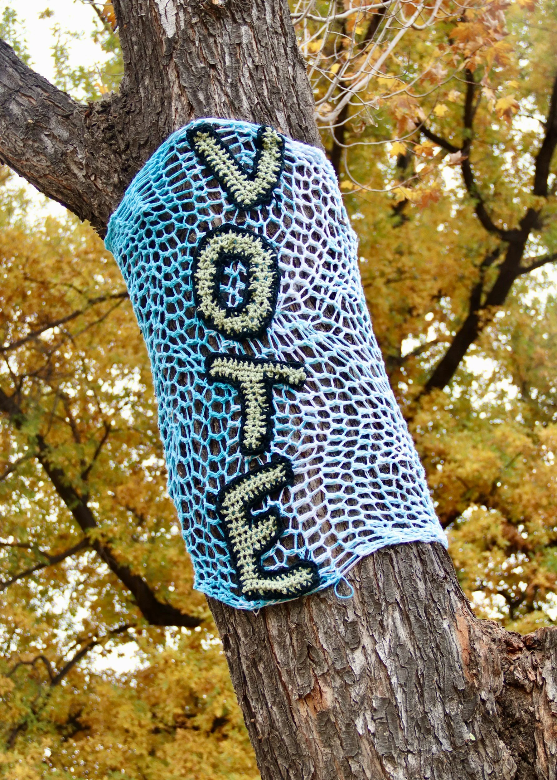 A crocheted yarn bombing sign with the word 'VOTE' on it, wrapped around a tree in an autumn park