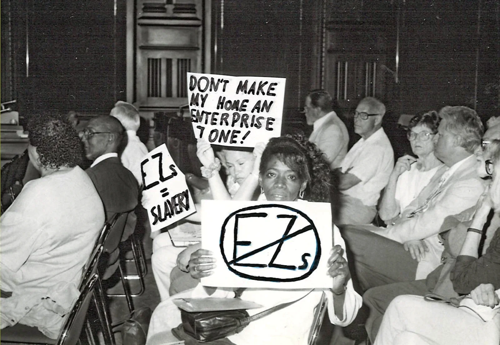 Black woman holding a "NO EZ's" sign at a public hearing
