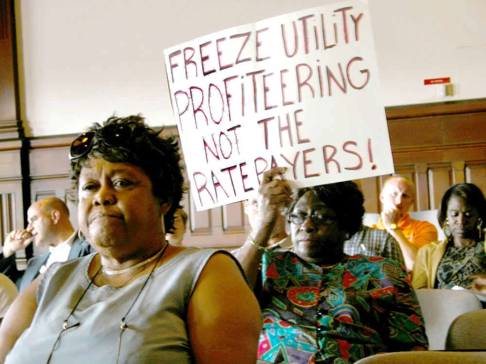 Five people attending a utility rate hearing, and one older black woman holding a sign saying "Frezz Utility Profiteering, not the ratepayers"