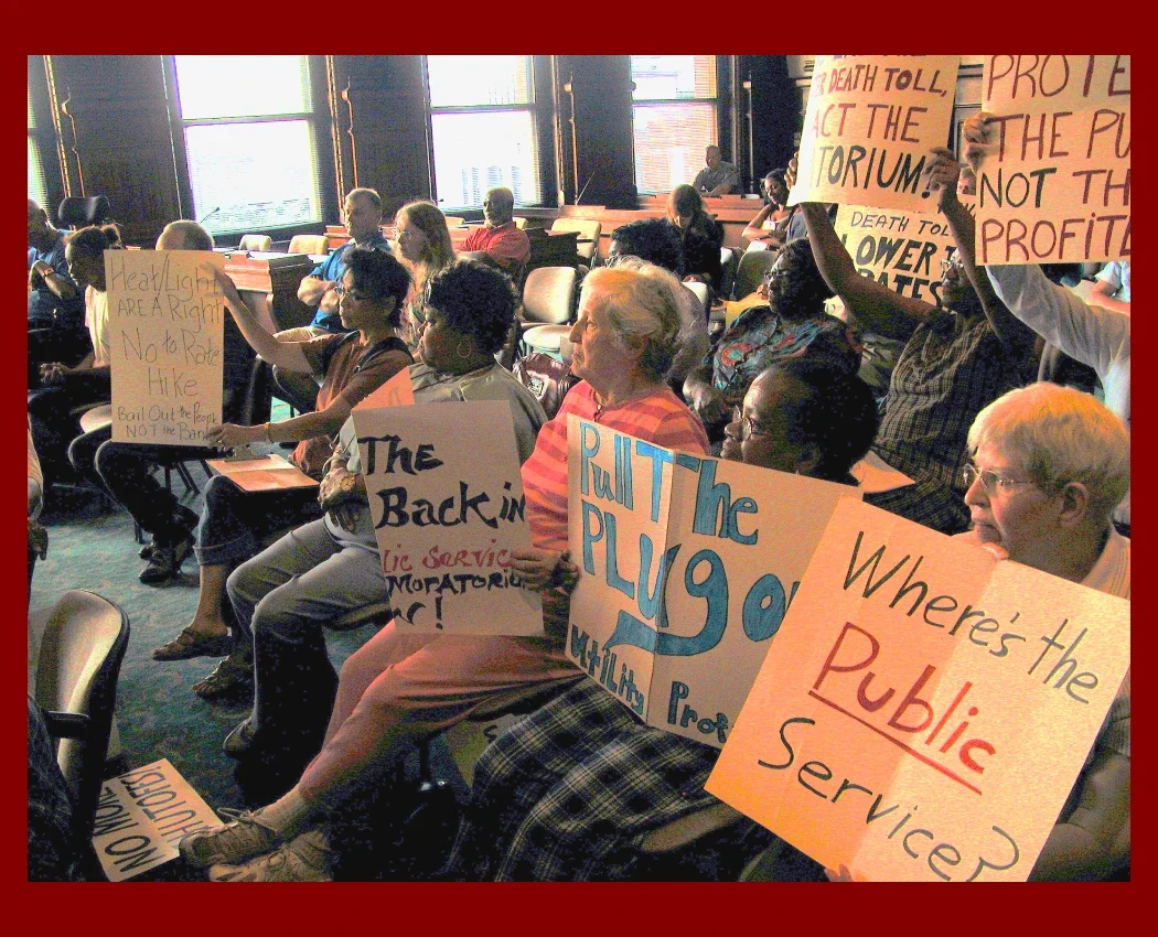 A group of more than 20 people in a hearing room carrying utility rate hike protest signs