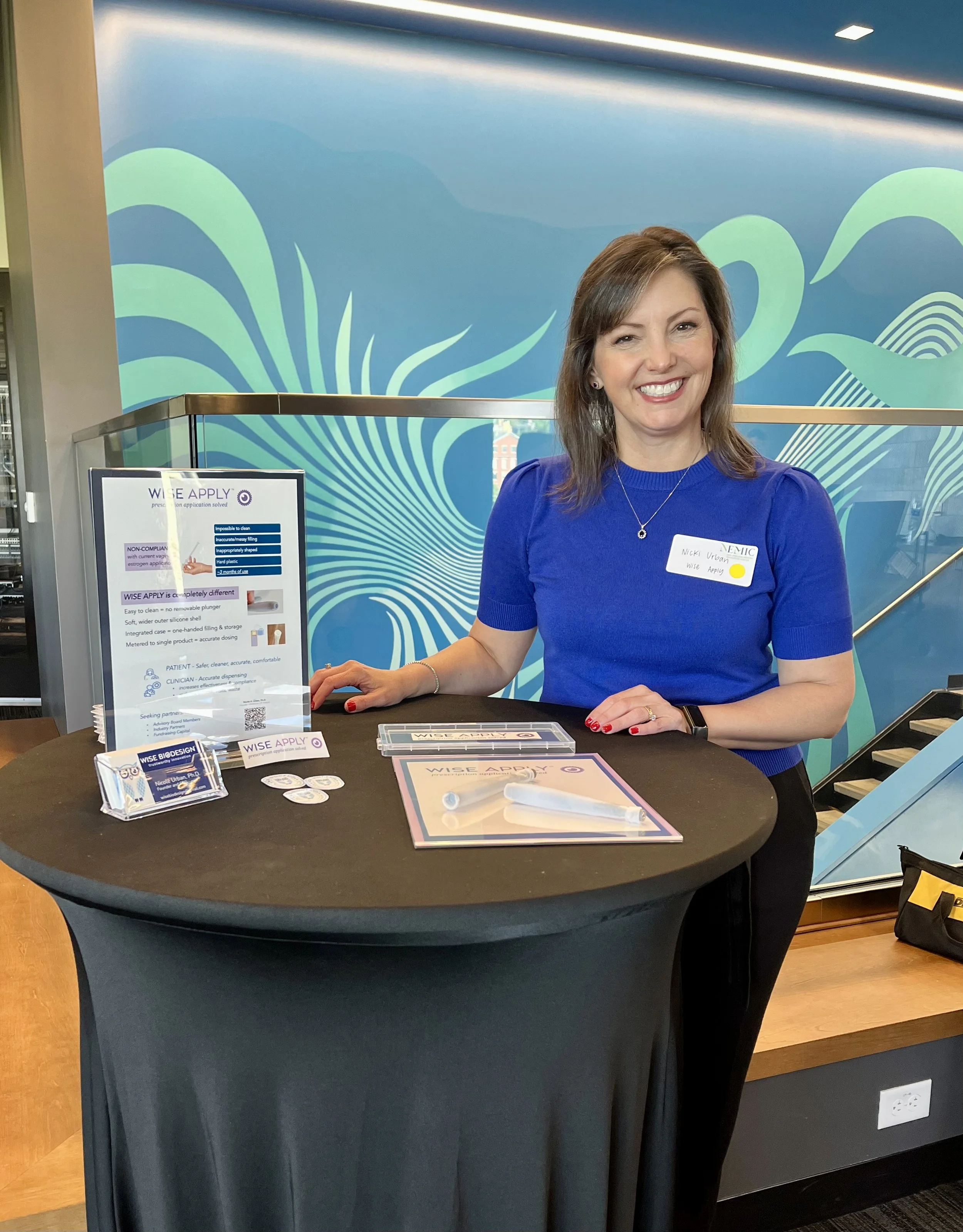 A woman with brown hair and a blue shirt standing behind a black round table with a display stand and some informational materials on it, in front of a blue abstract wall with wave patterns, inside a modern building.
