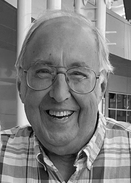 A smiling elderly man with glasses, wearing a plaid shirt, in an indoor setting with modern architecture in the background.