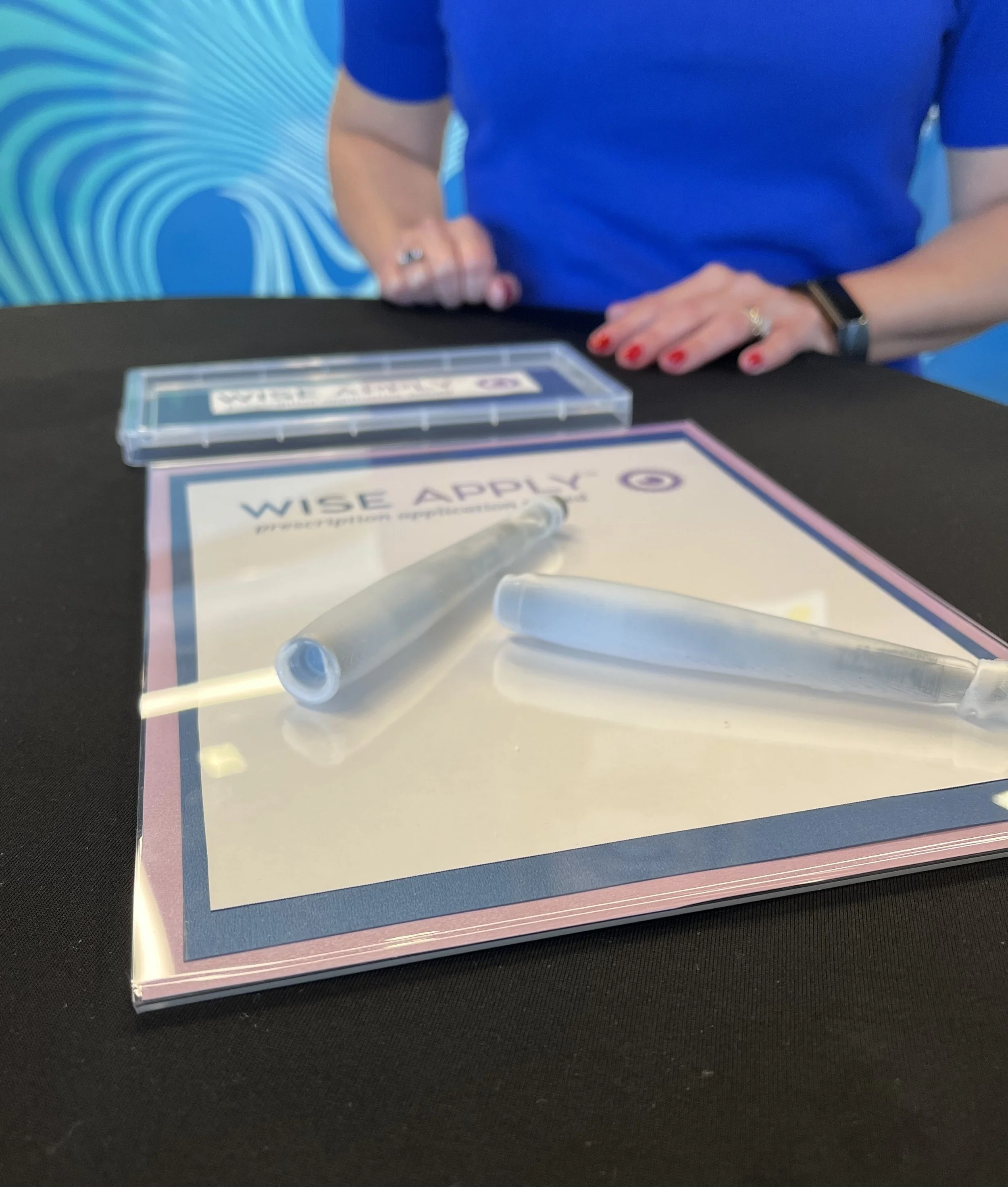 Close-up of a person with red painted nails and a watch, sitting at a table with a blue background featuring a swirling pattern, a certificate partially visible, and medical supplies including a syringe and a small clear box with labels.