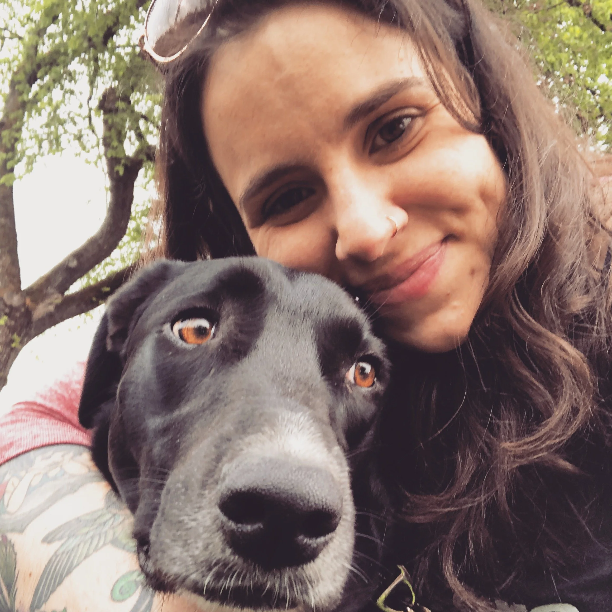 A person with long wave hair and a nose ring smiling and hugging a black dog with brown eyes outdoors near a tree.