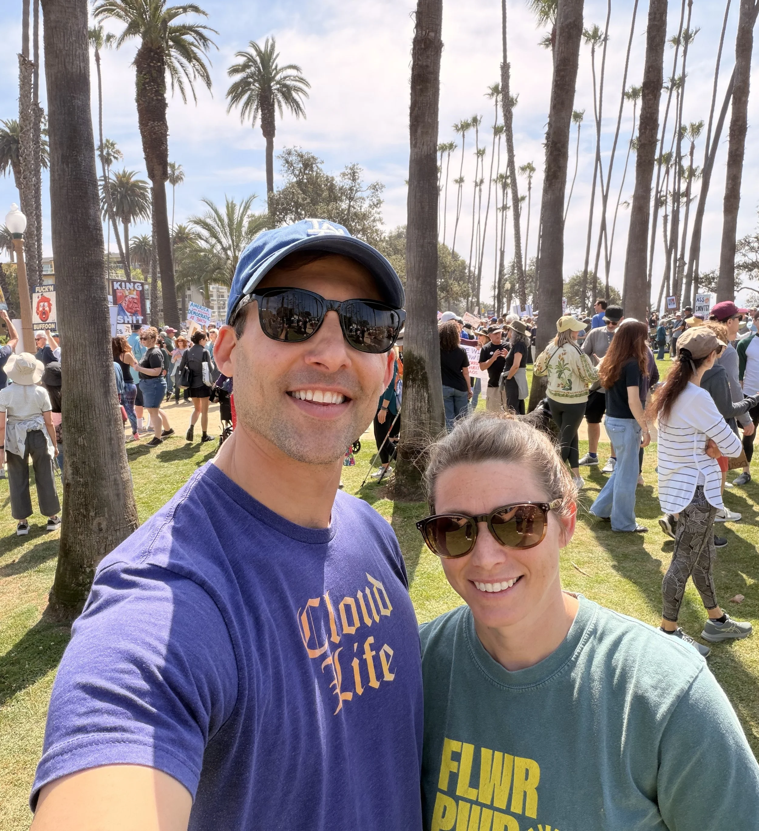 Harry Leshner and Hana Leshner taking a selfie at a protest or rally in a park with tall palm trees and a crowd of people holding signs in the background.