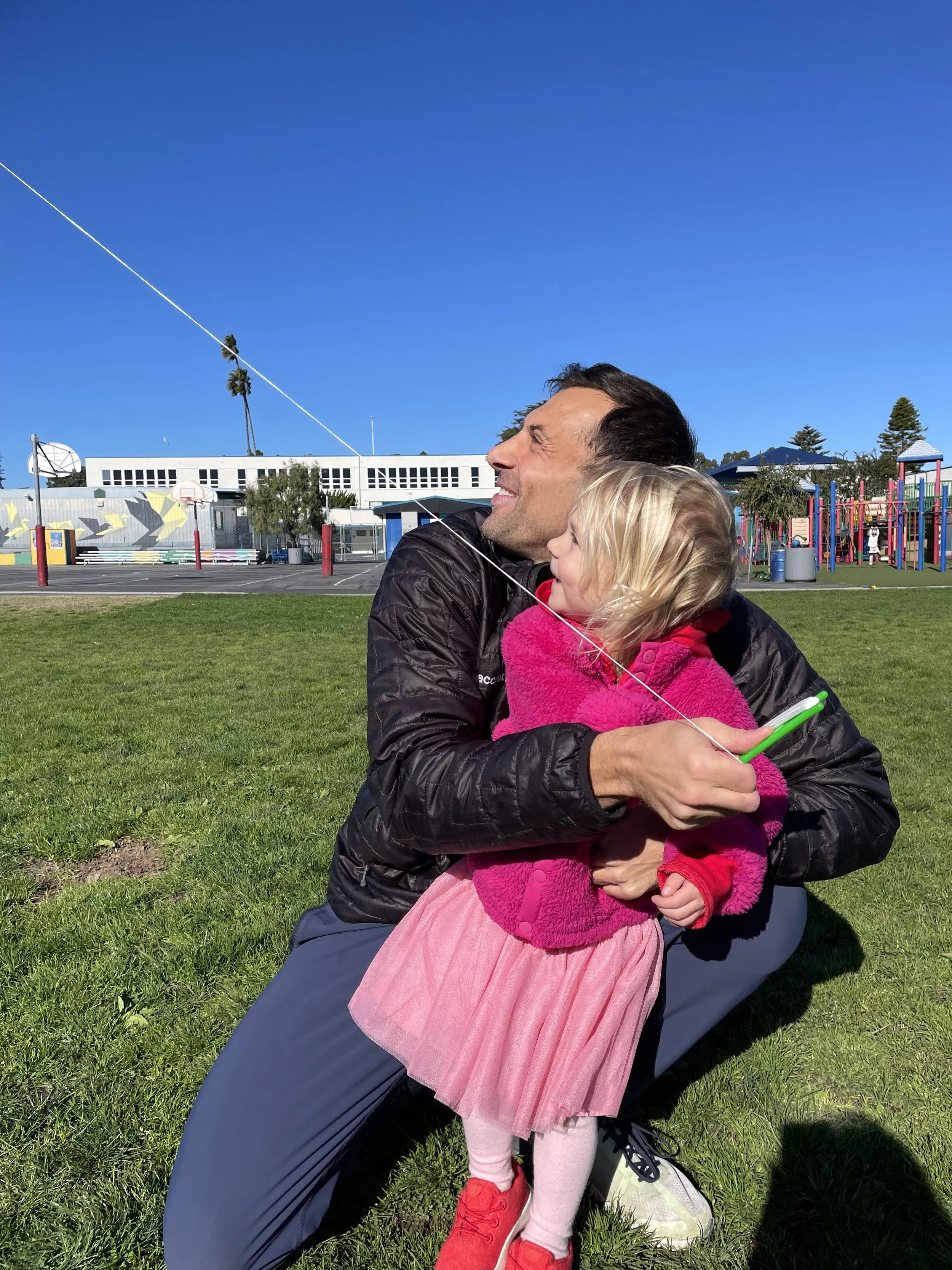 Harry Leshner and his young daughter sit on the grass outdoors, flying a kite against a clear blue sky. The girl is dressed in a pink dress and fuzzy pink vest, holding a green kite handle, while the man holds her and smiles.
