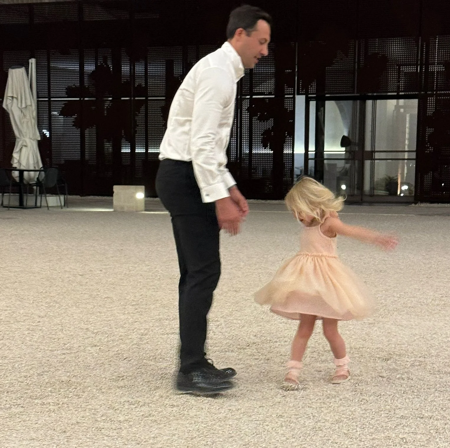 Harry Leshner in black pants and a white shirt is dancing with his young daughter in a pink dress.