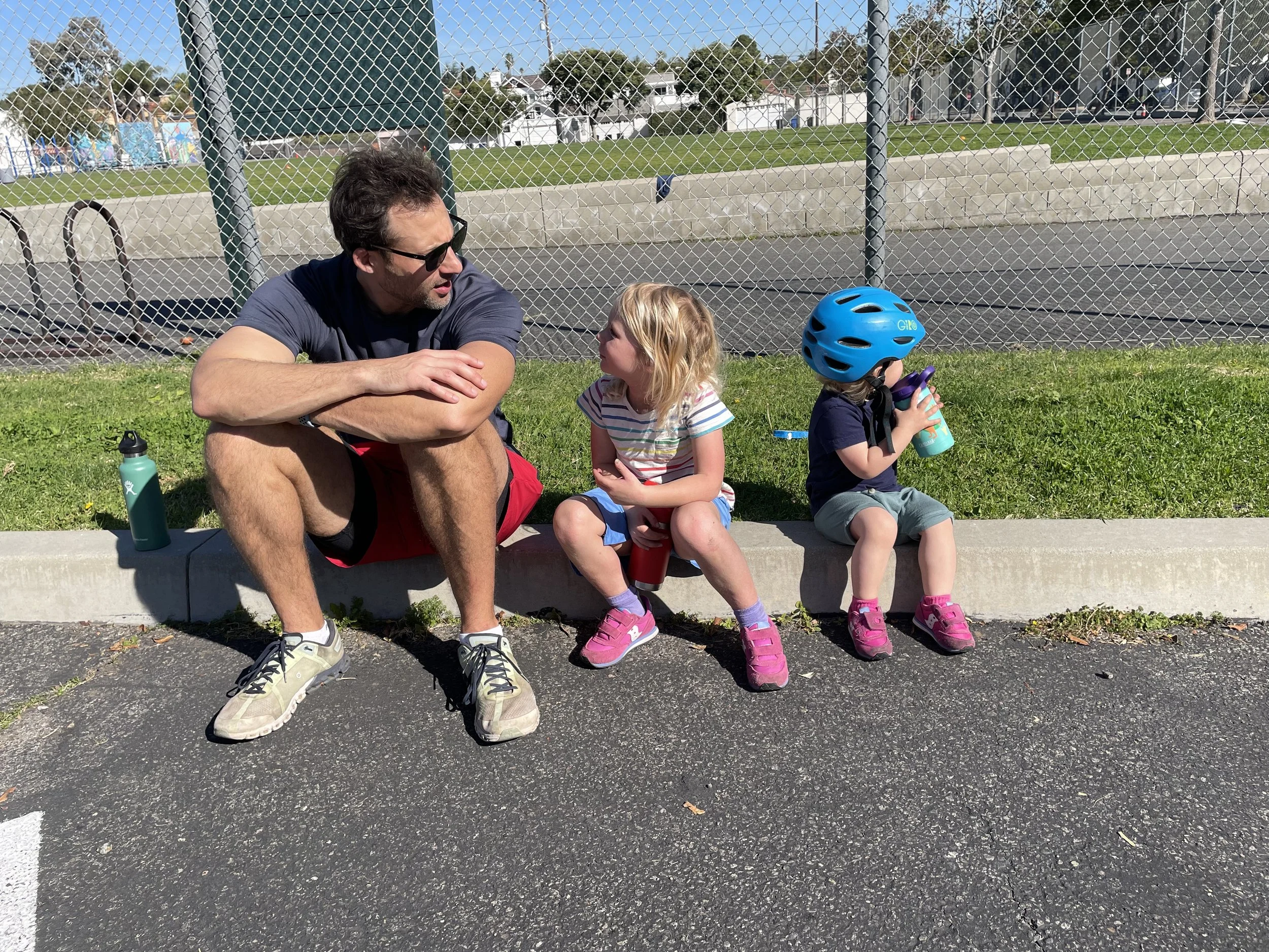 A man with sunglasses talking to two young girls sitting on a curb at a park. One girl wears a bicycle helmet and drinks from a cup, the other girl watches, with water bottles nearby. A chain-link fence and grassy field are in the background.