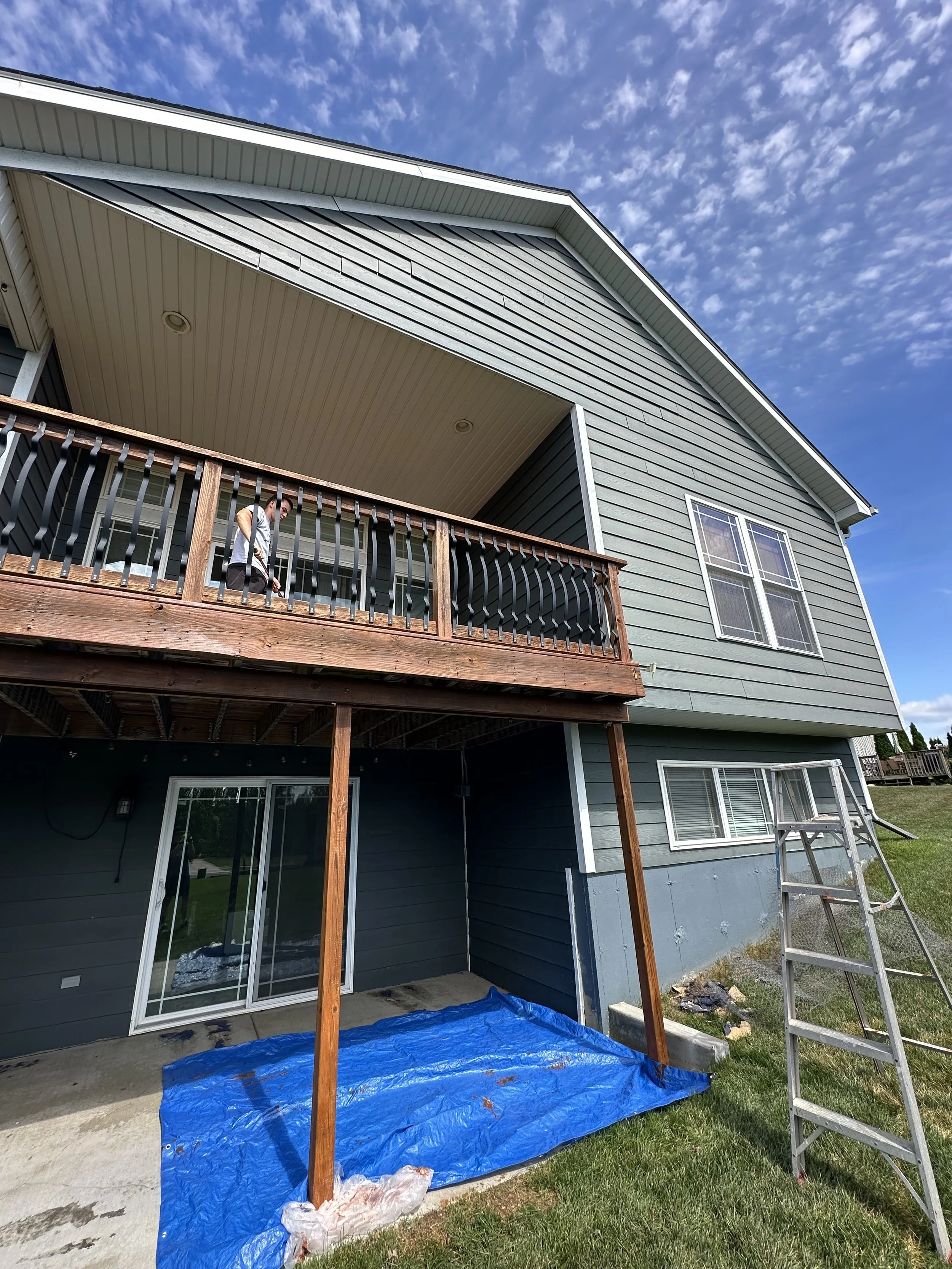 A two-story house with a blue exterior and a wooden balcony. A person on the balcony is working, and the area below the balcony is covered with a blue tarp. A ladder is leaning against the house, and a slide is nearby.