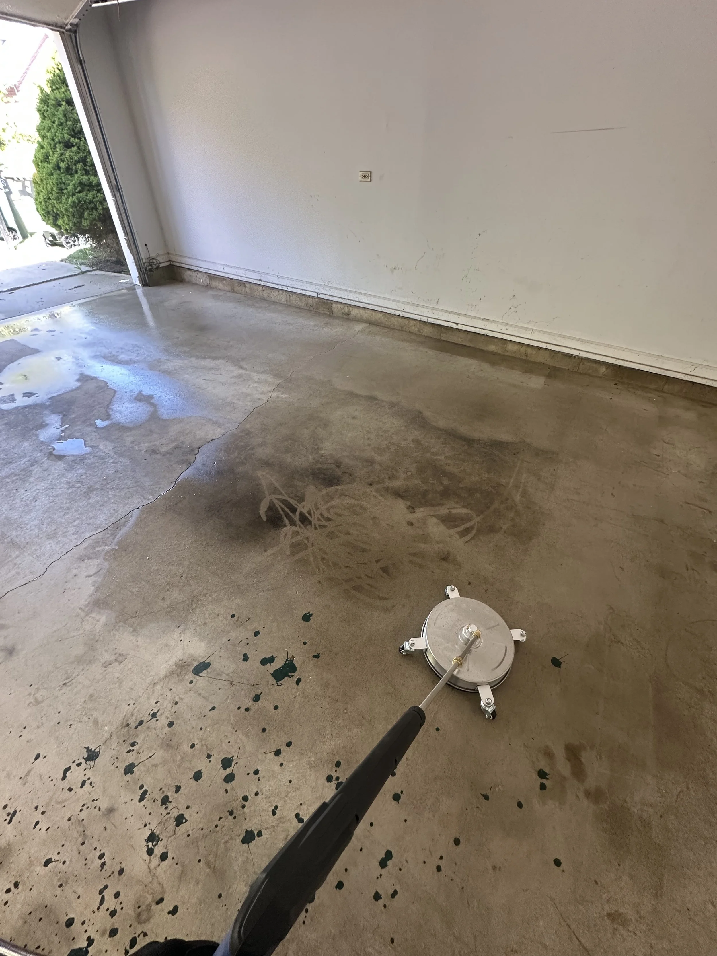 A clean concrete garage floor with a power washer cleaning dark stains, with a side door open showing outdoor greenery.