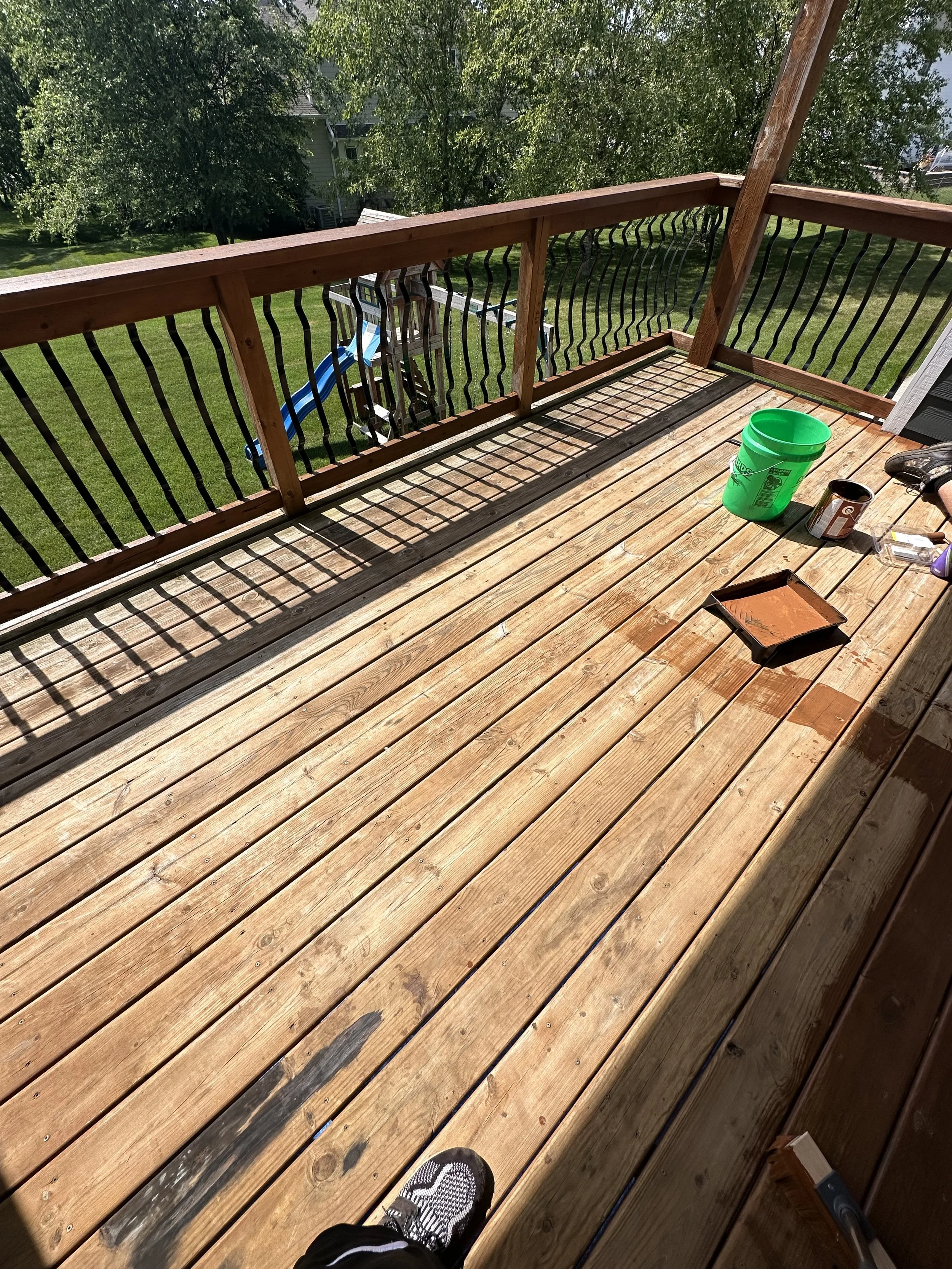A wooden deck with some tools and paint supplies, including a green bucket, a paint can, and a paint tray, with a grassy backyard and trees in the background.