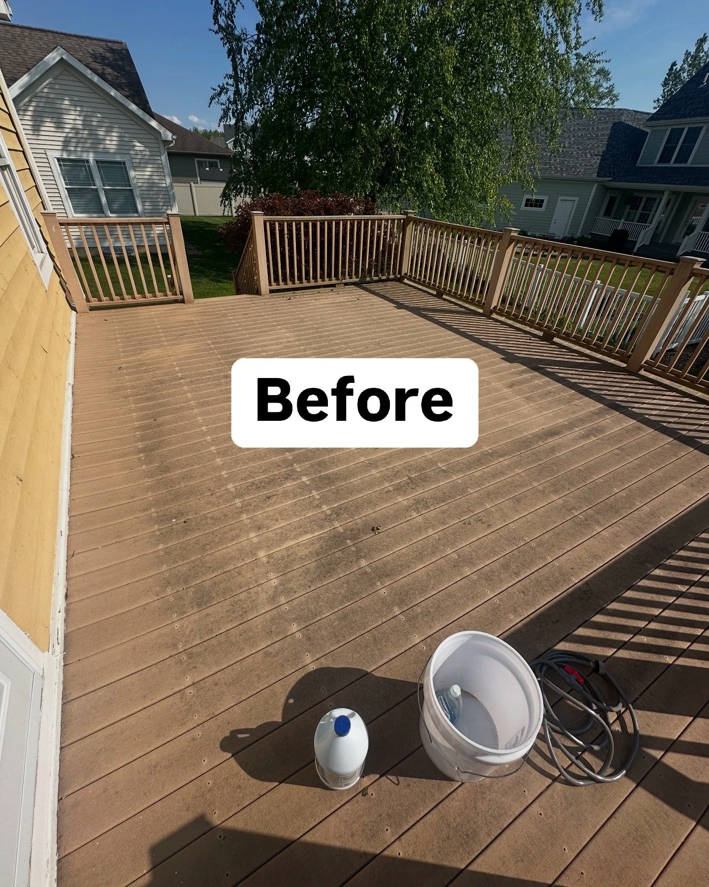 A wooden deck with cleaning supplies, including a bucket, a bottle, and a hose. The deck is dirty and has dust and debris on it, with houses and trees visible in the background.