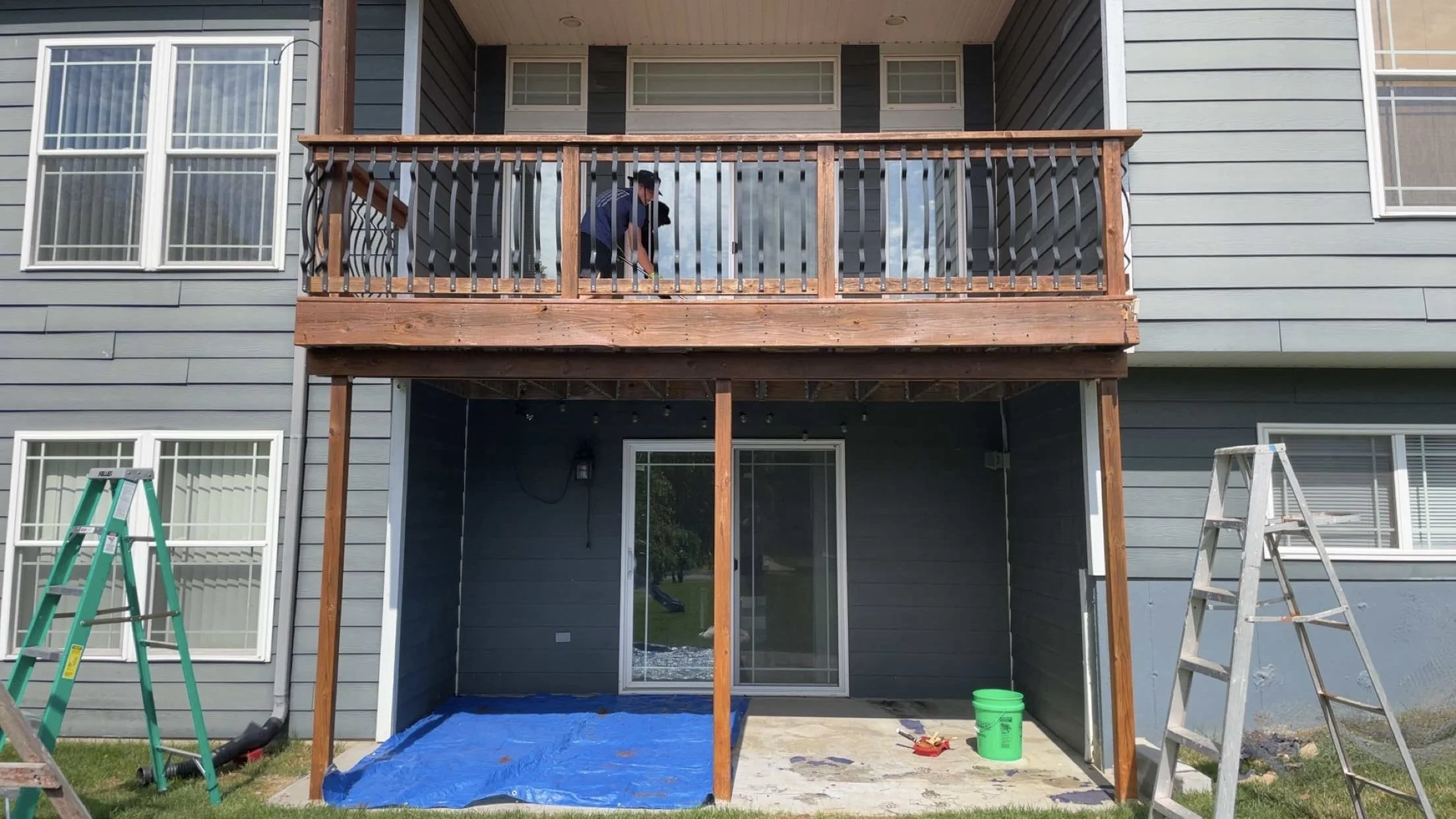 A person on the upper deck of a two-story house painting or staining the wood railings, with ladders and tools on the porch below, as part of a home renovation.