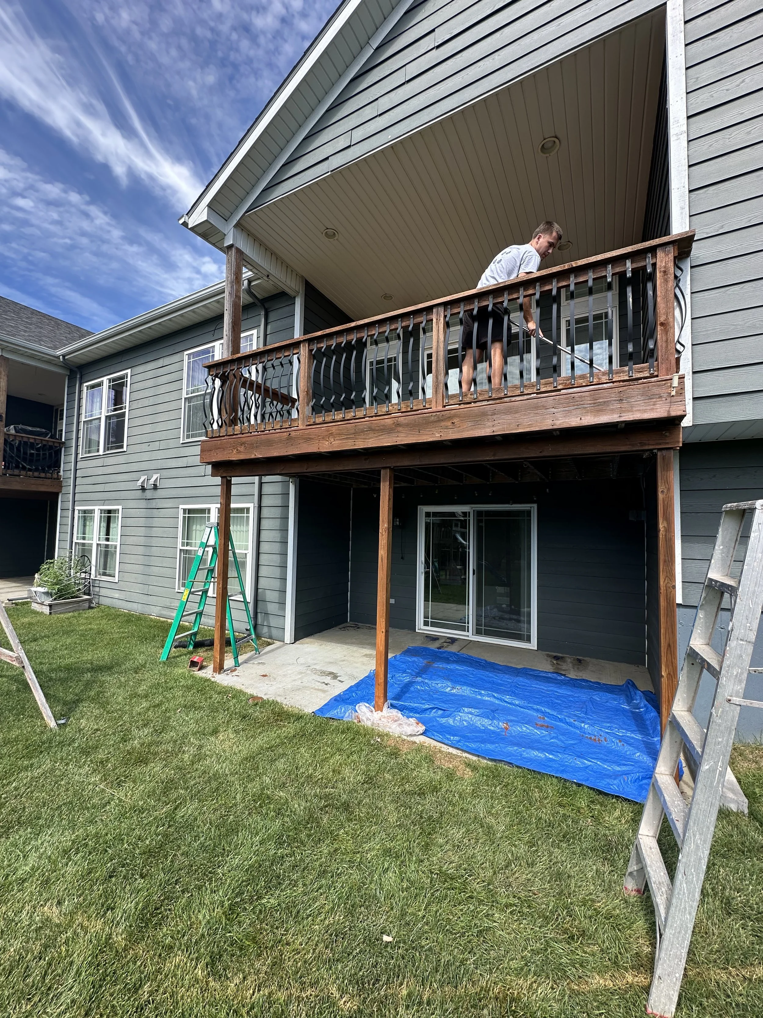 A person working on a wooden balcony of a multi-story house, painting or staining the railing, with ladders and a tarp on the ground, under a partly cloudy sky.
