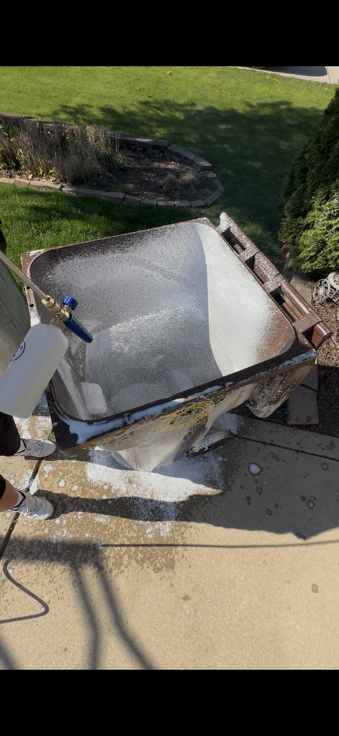 Person using a foam cannon to clean a trash bin outside, with soap suds overflowing and covering part of the sidewalk.