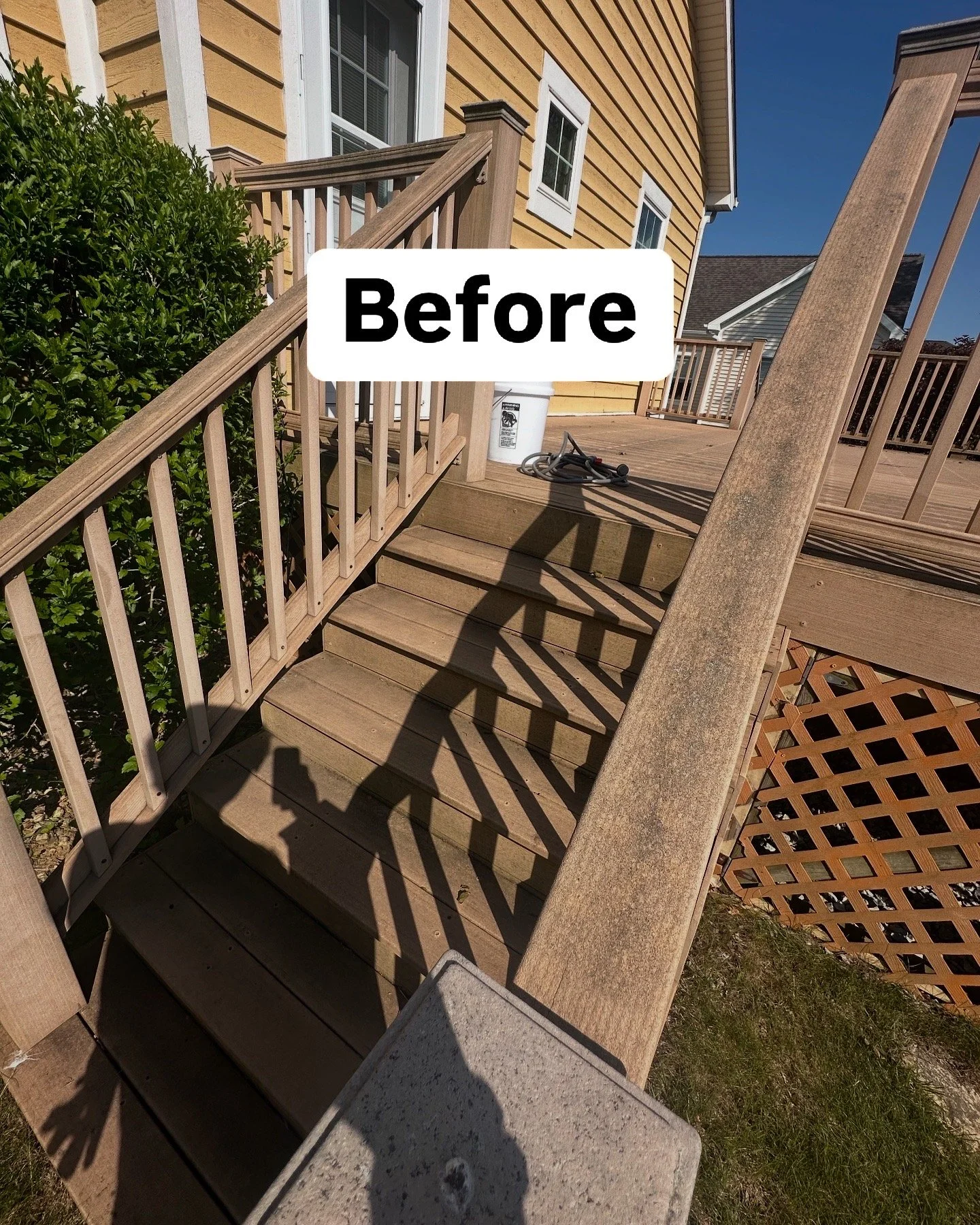 Backyard wooden deck with stairs, railing, and shadow patterns, yellow house with white trim, gardening tools, and greenery.