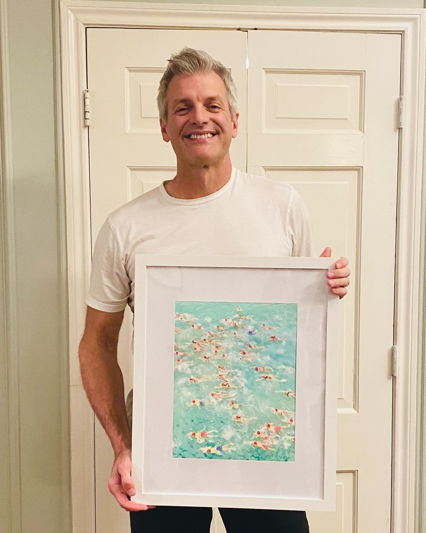A man smiling and holding a framed painting of a crowded swimming pool filled with swimmers.