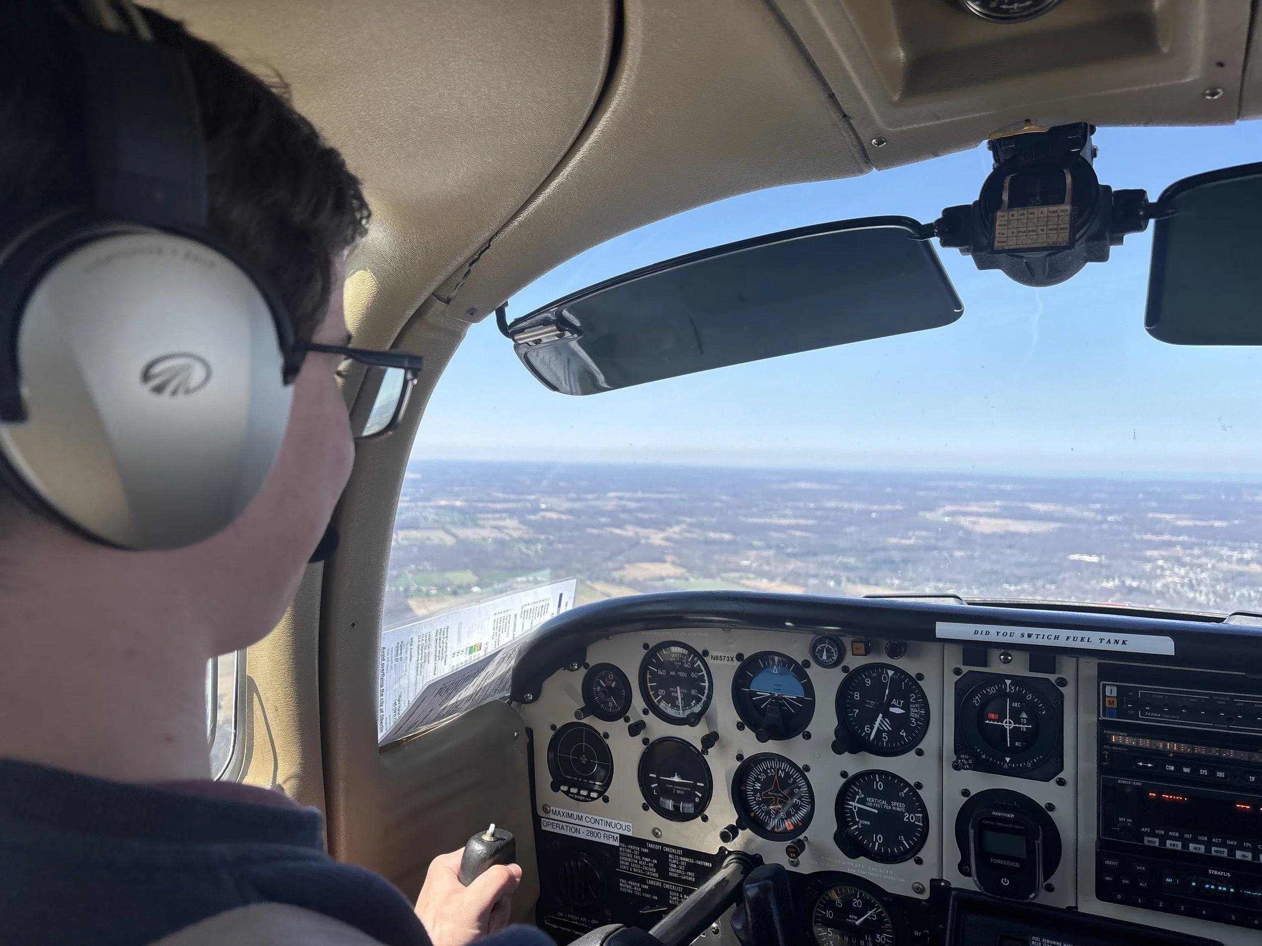 A pilot flying an aircraft seen from inside the cockpit, with cockpit instruments and controls visible, and a landscape of fields and sky outside the window.