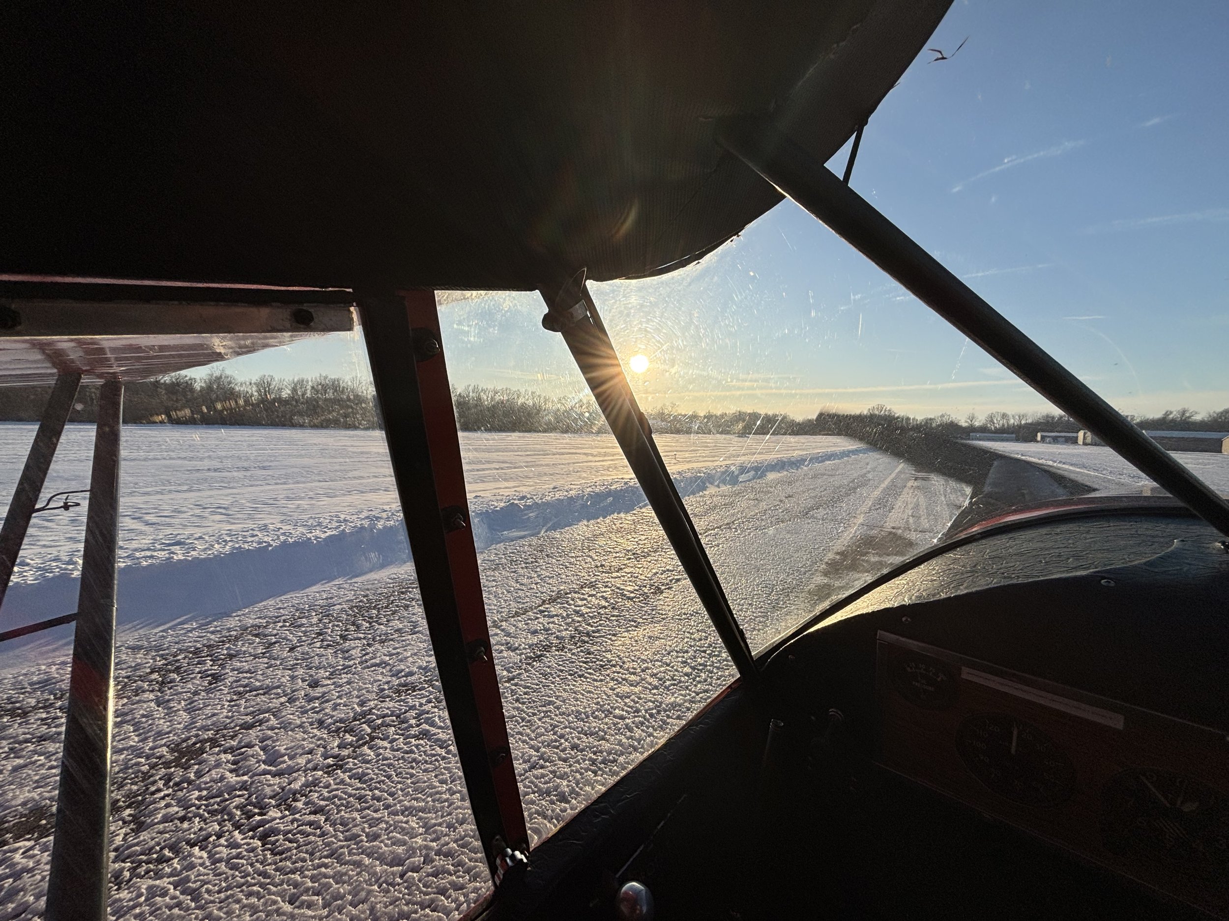 View from inside a small aircraft, looking out over a snowy landscape with the sun low in the sky and a clear blue sky. Part of the aircraft's wing and support struts are visible.