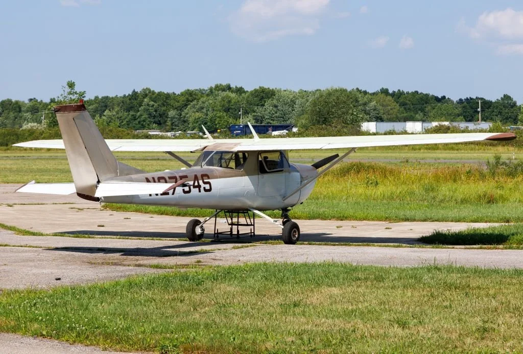 Small white airplane with tail number N27545 parked on a tarmac runway, with grassy areas and trees in the background under a partly cloudy sky.