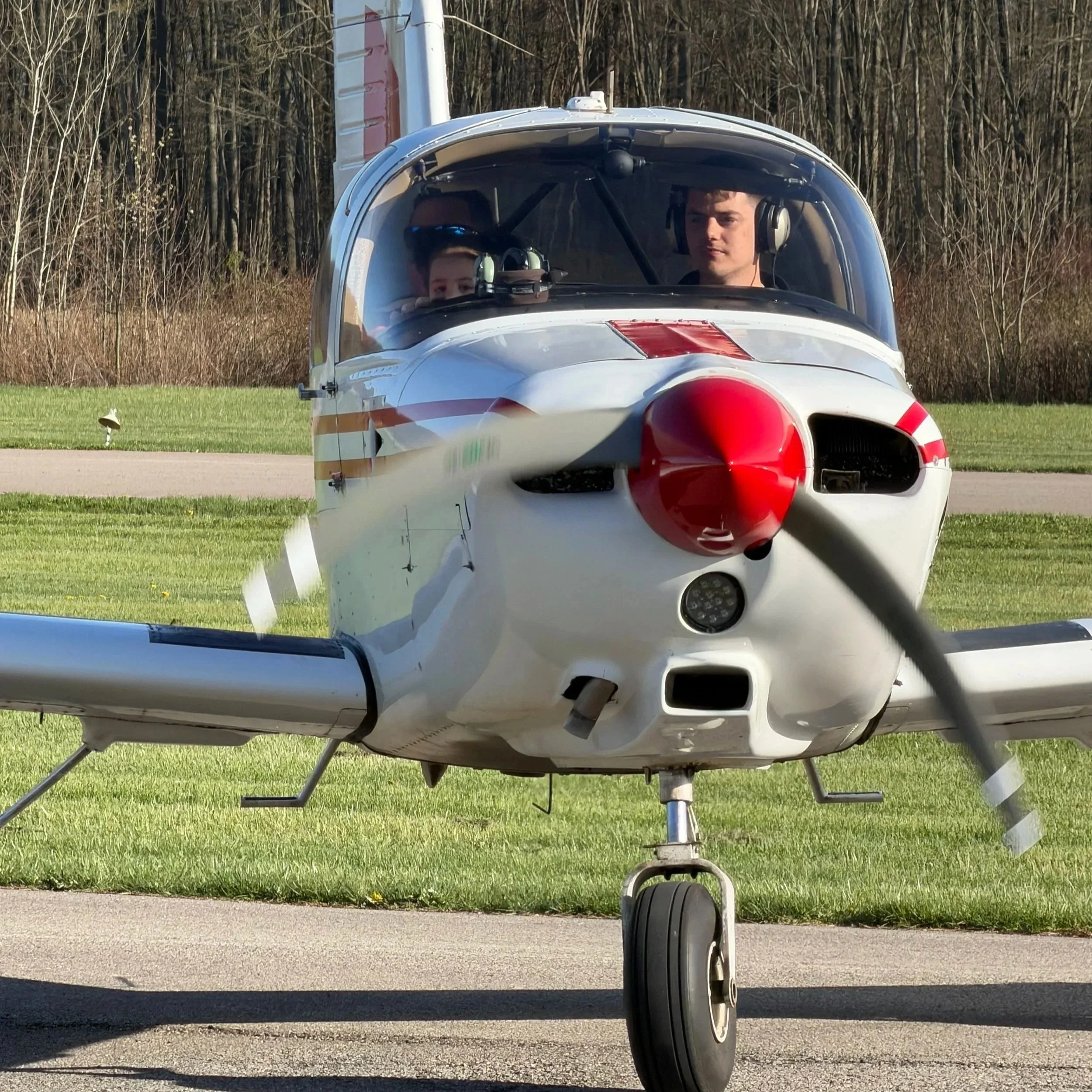 A small helicopter with a white and red fuselage is on the ground, with a person inside the cockpit wearing headphones and looking ahead, set against a backdrop of trees and clear sky.