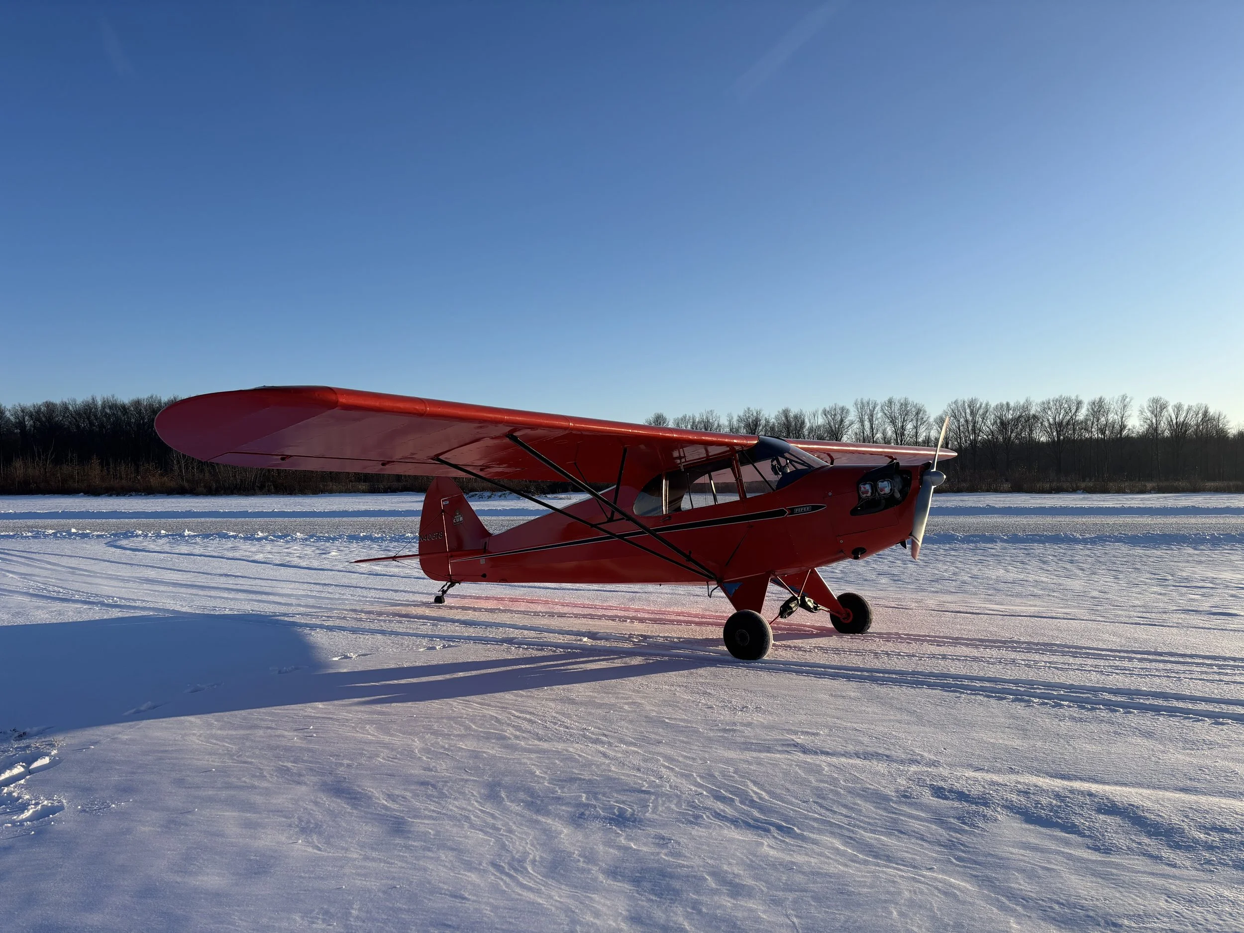 A red small airplane parked on a snow-covered field under a clear blue sky with a line of trees in the background.