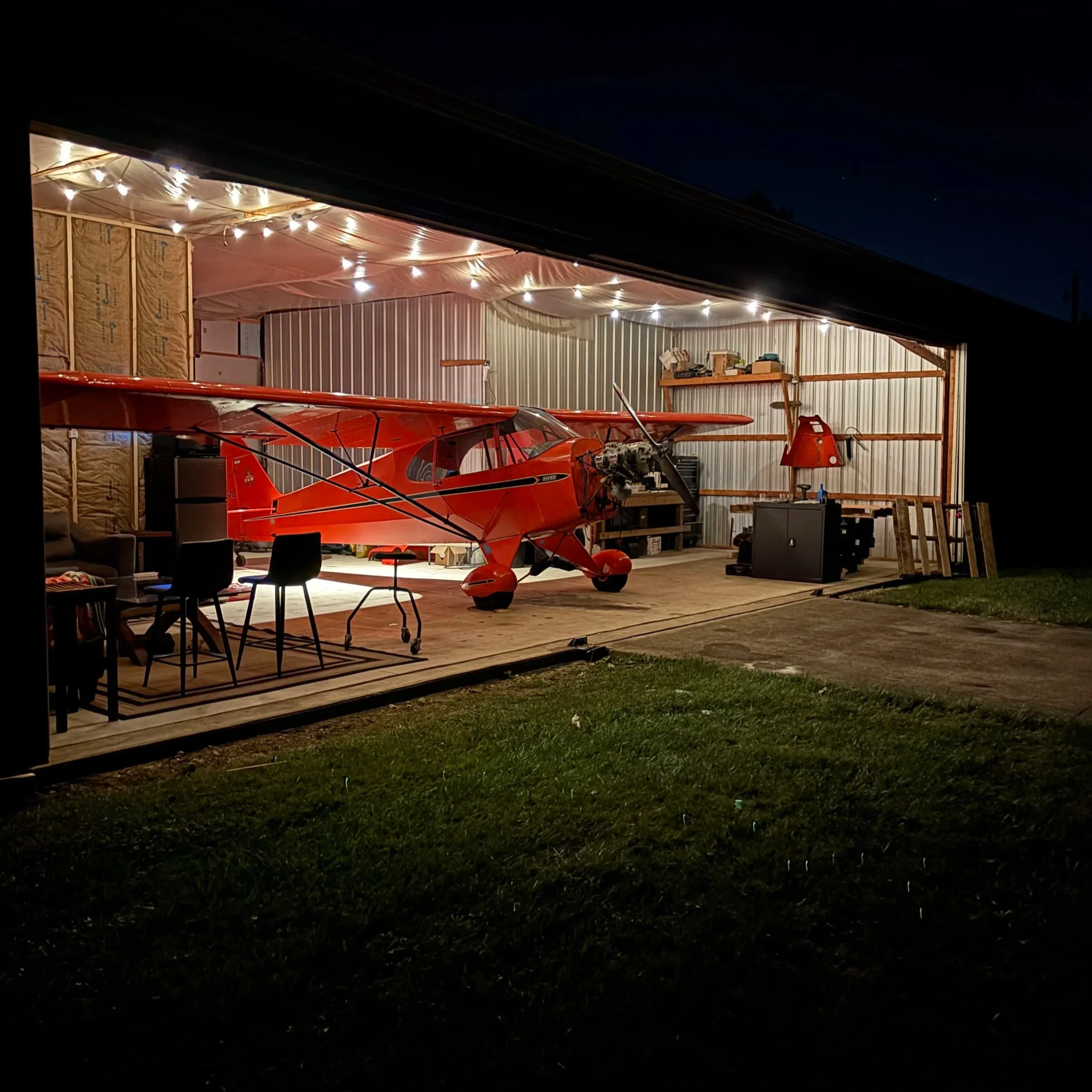 A red aircraft is inside a hangar with string lights hanging from the ceiling, and the hangar has an open side revealing grasses and night sky outside.
